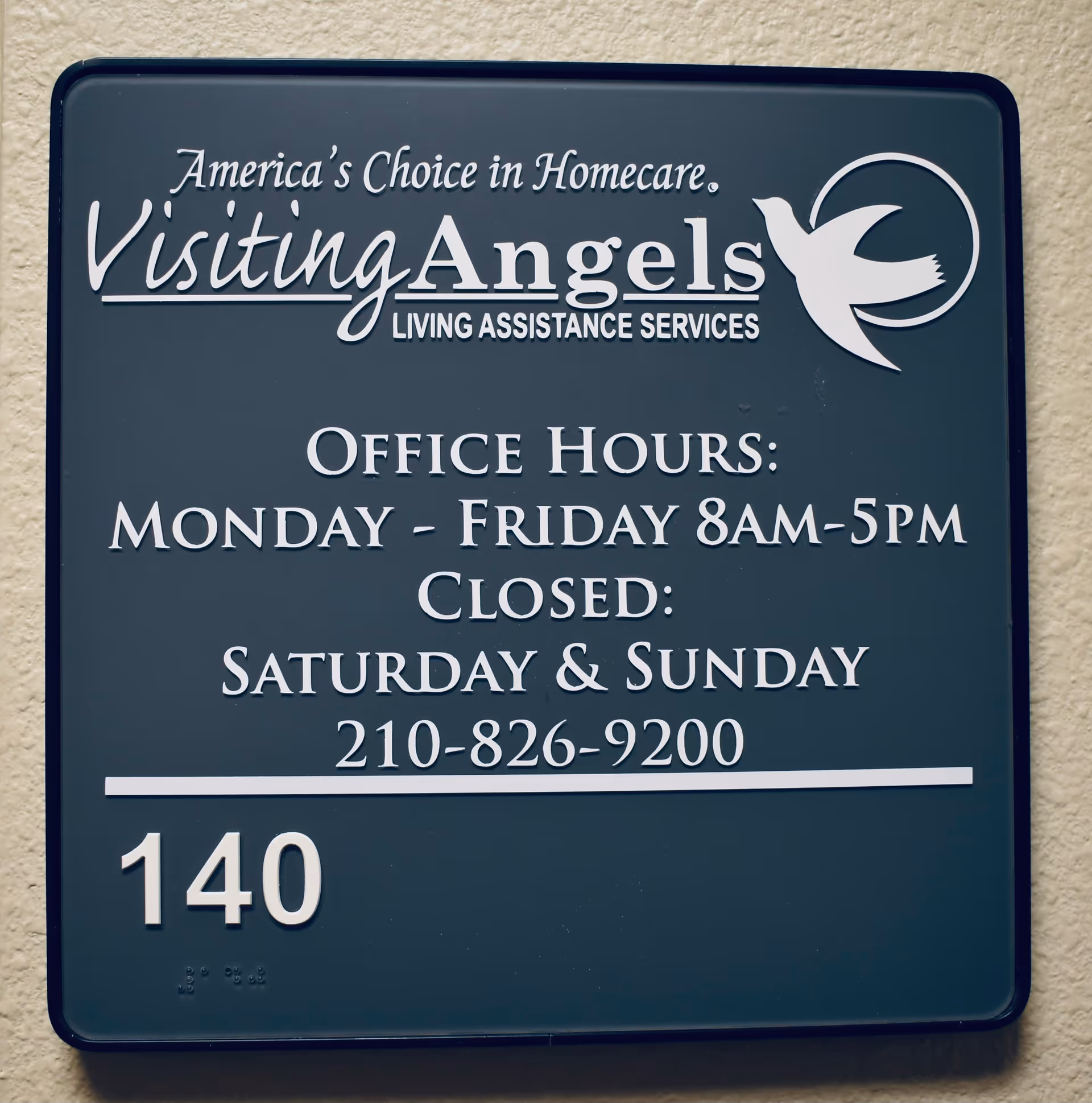 Blue rectangular sign on beige wall displaying Visiting Angels office hours and contact.