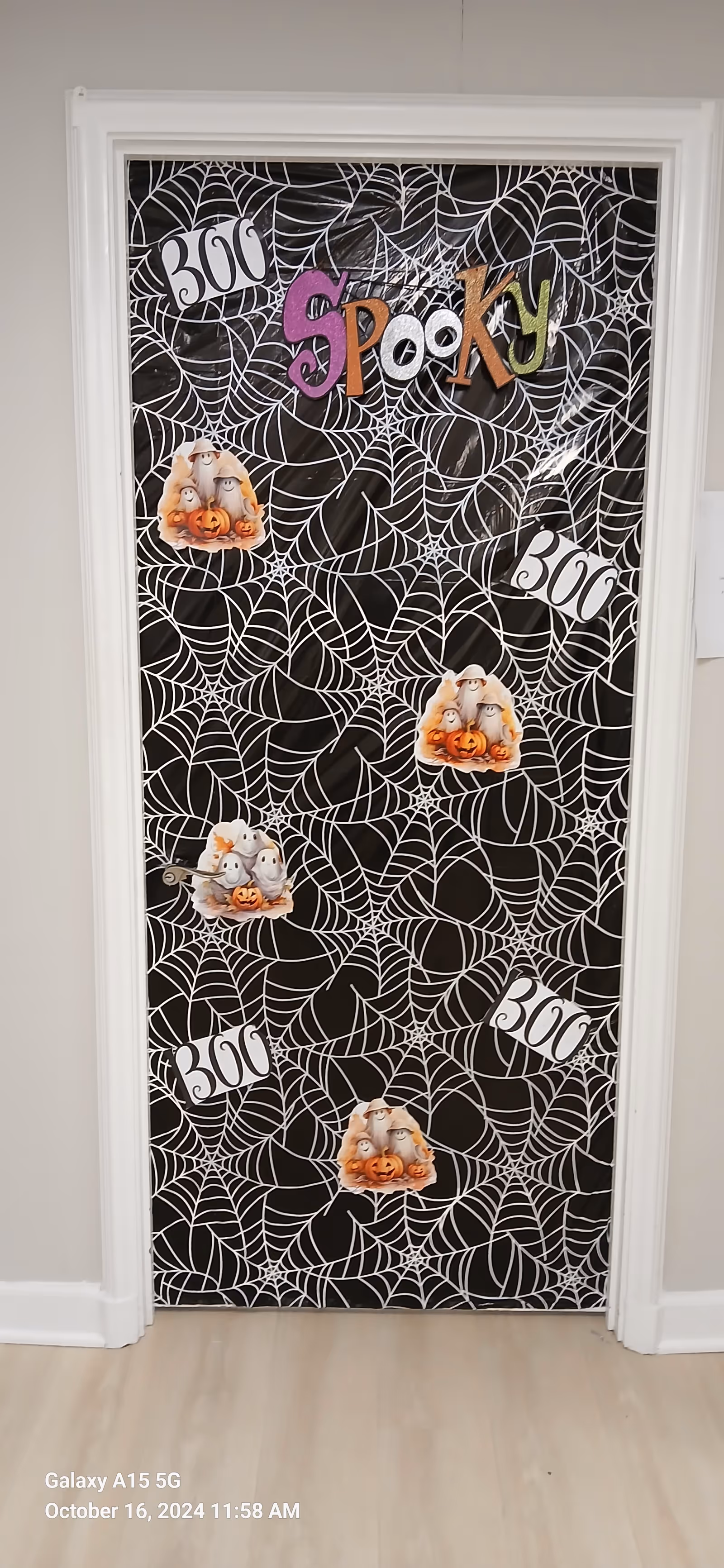 Halloween-themed door decoration with spider webs, pumpkins, and spooky lettering in a hallway.