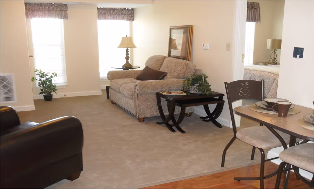 Living room in a senior facility with sofa, dining table, and windows.
