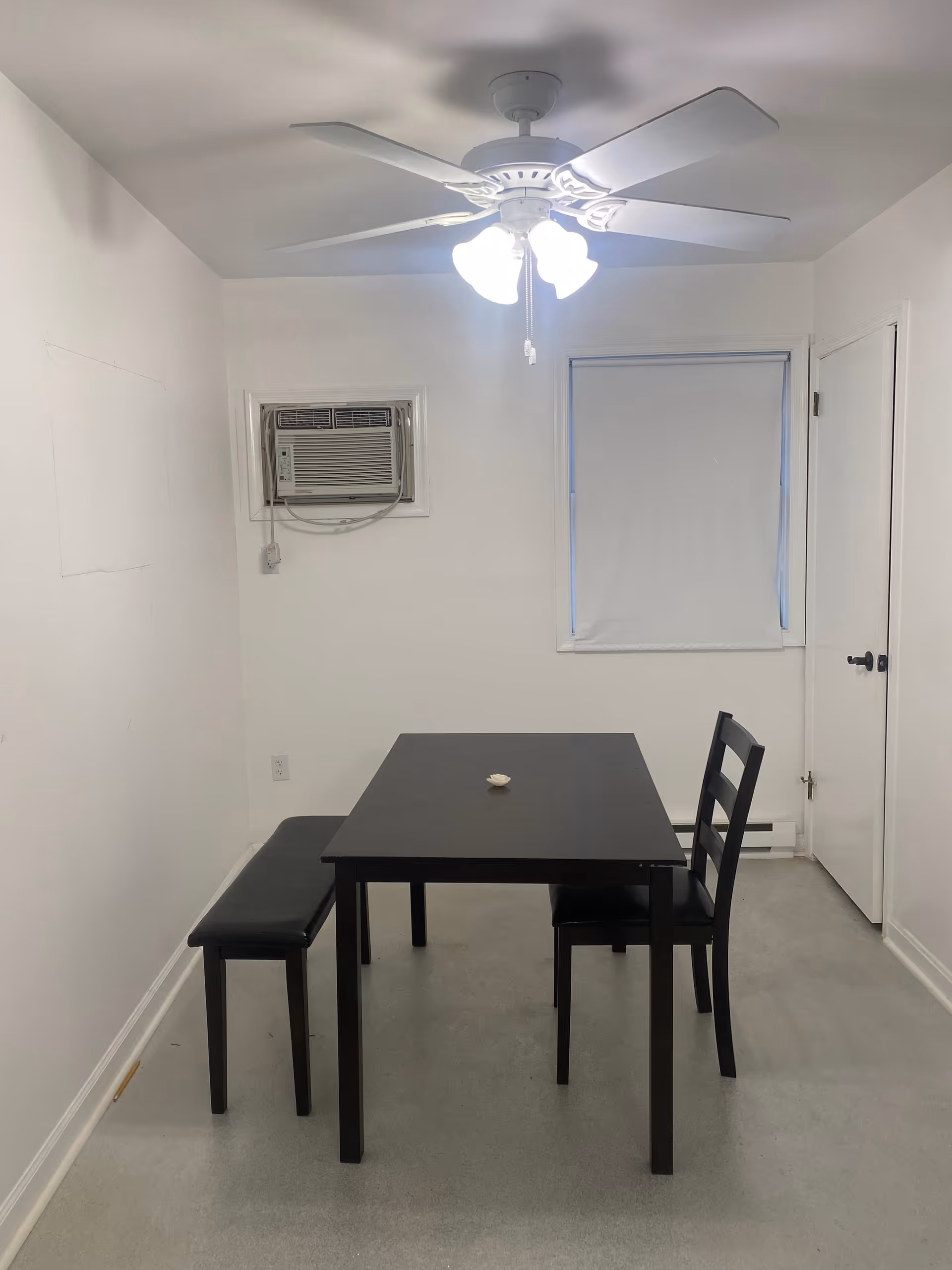 Small minimalist dining area with a dark table, bench, chair, ceiling fan, and window.