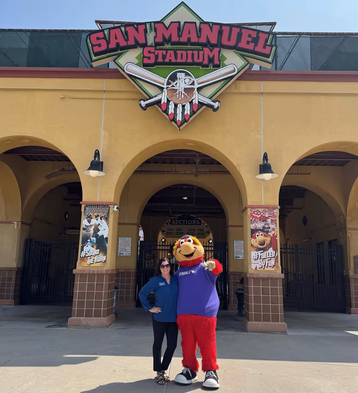 Two people pose with a mascot in front of San Manuel Stadium entrance.