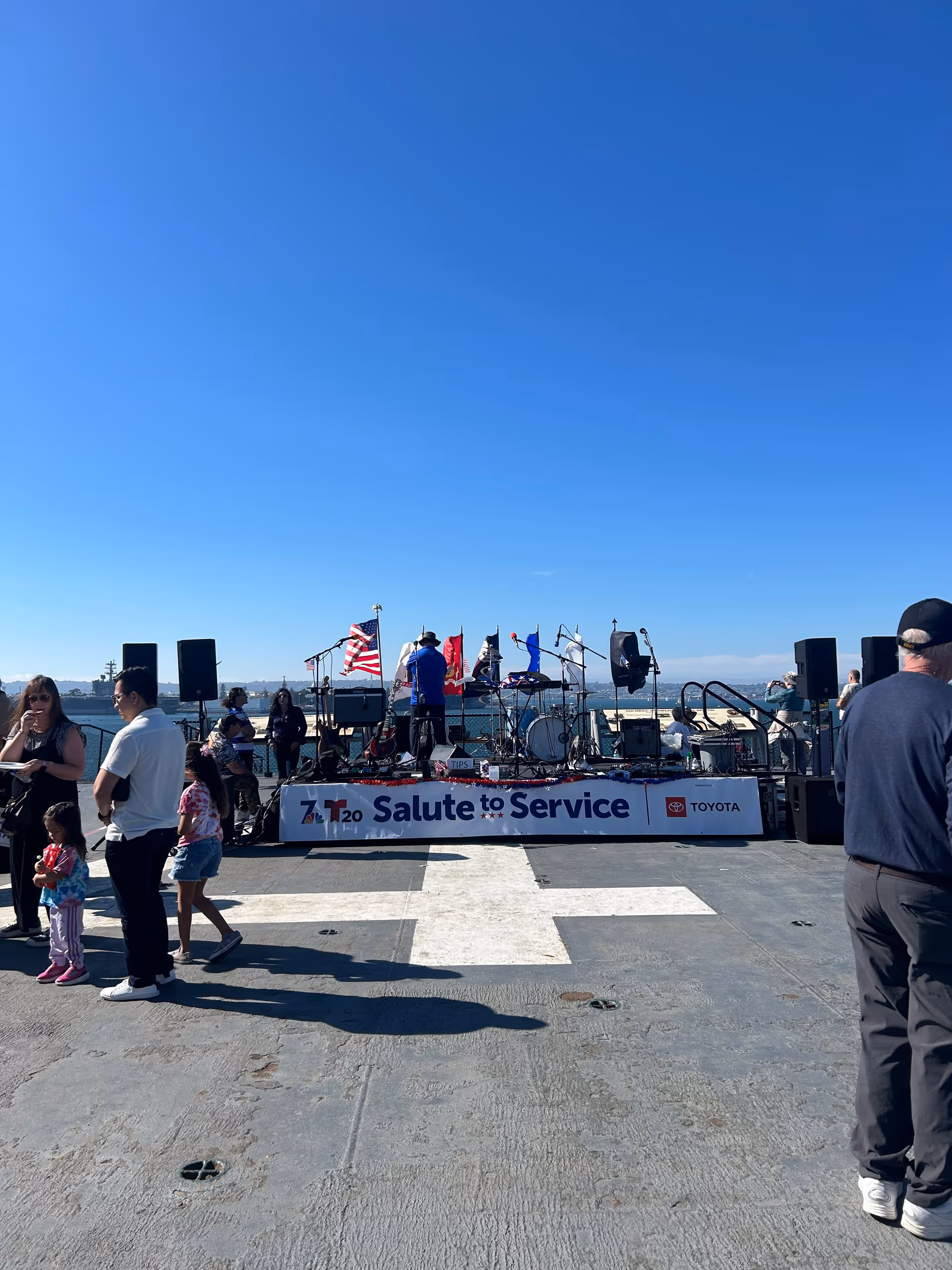 Outdoor waterfront stage with a band performing and spectators gathered nearby.