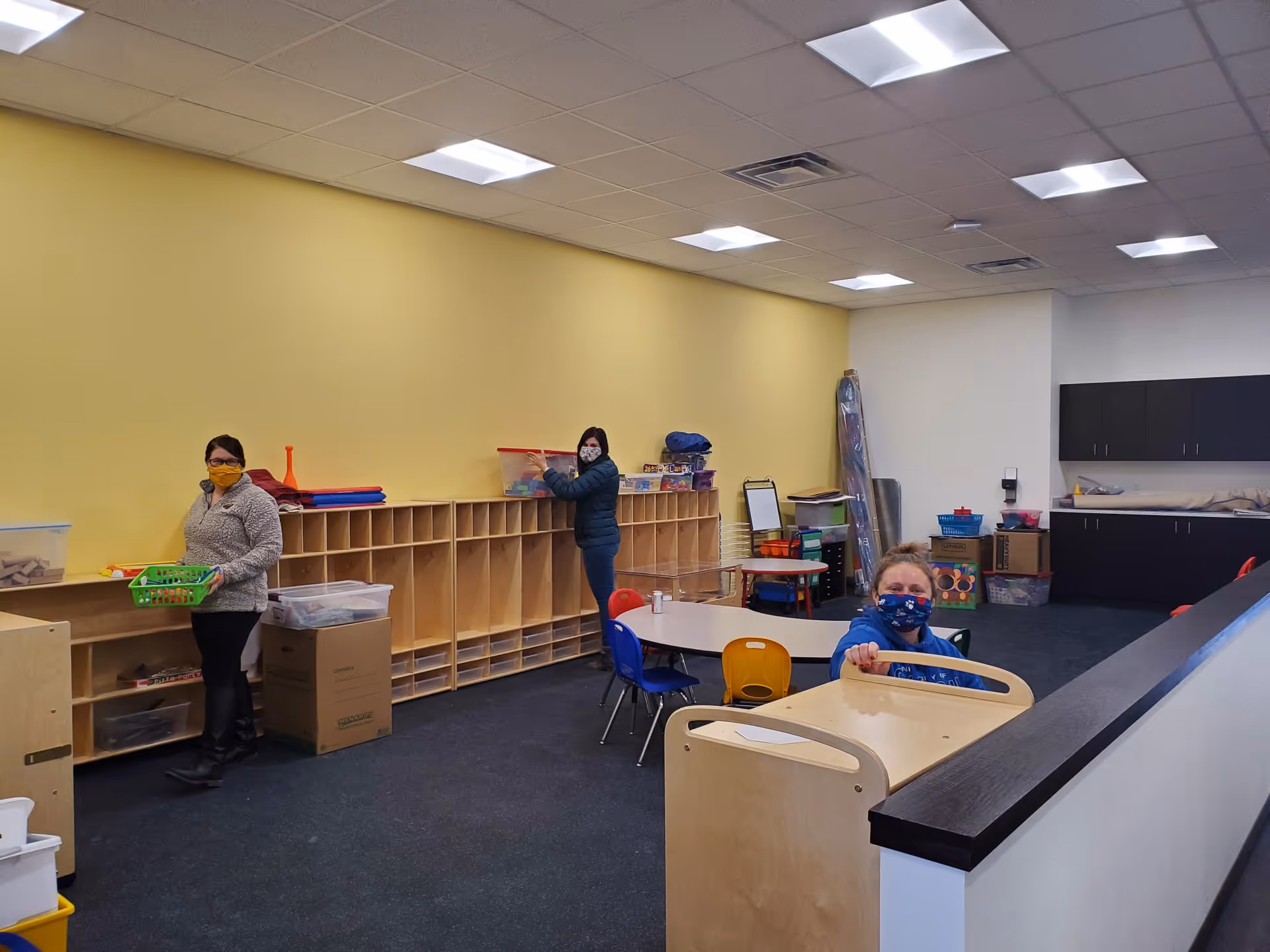 Bright classroom and activity room with shelves, tables, and three masked people.