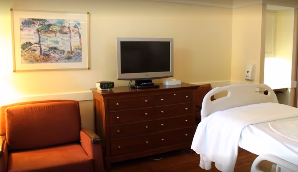 Hospital-style patient room with bed, dresser, TV, and orange chair.