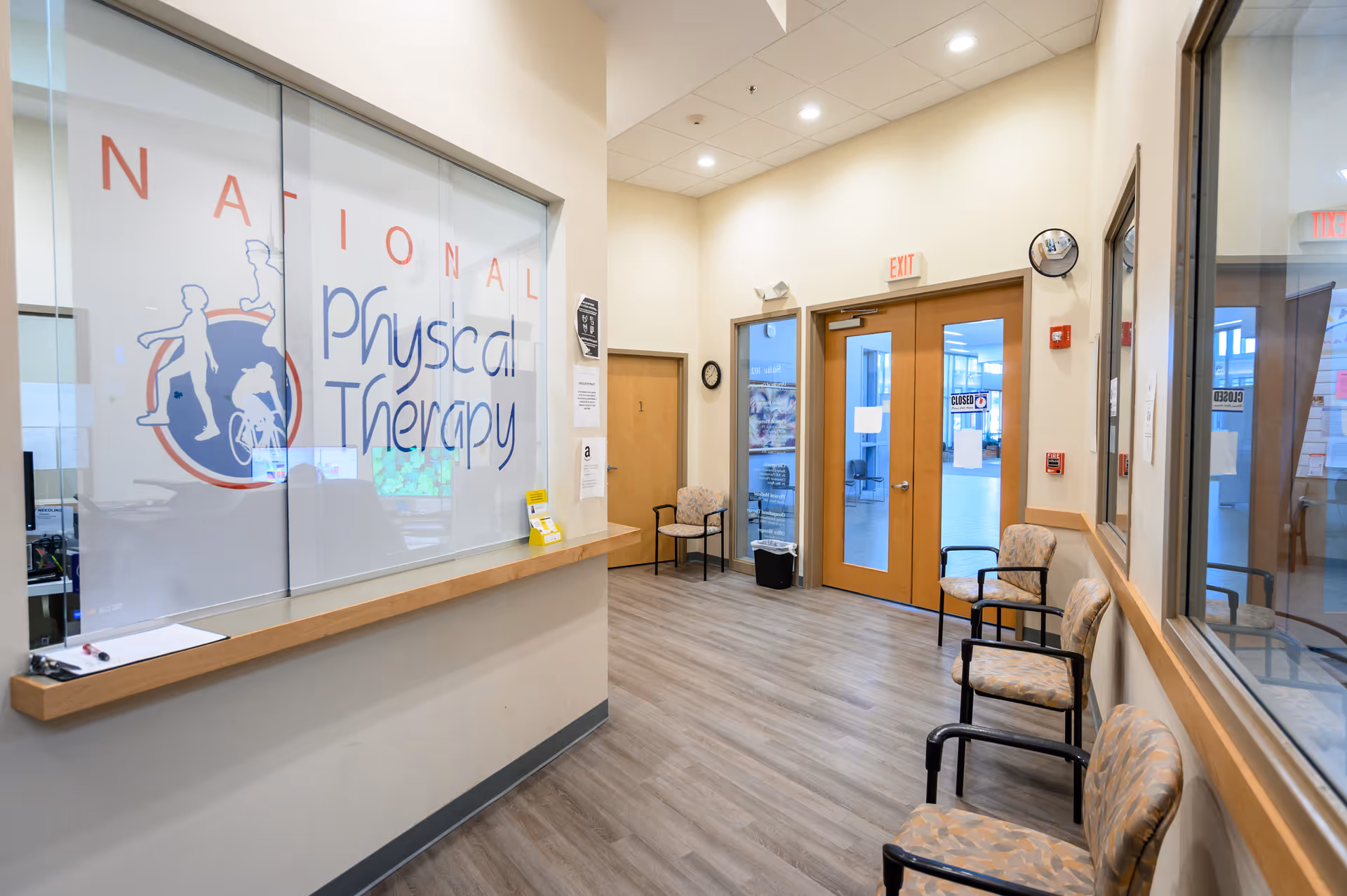 Bright clinic reception area with glass wall signage, a wooden counter, and seating along the hallway.