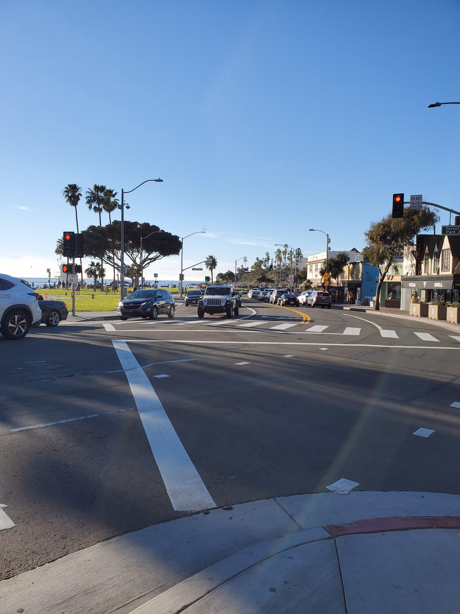 Sunlit coastal street intersection with cars, palm trees, and seaside shops.
