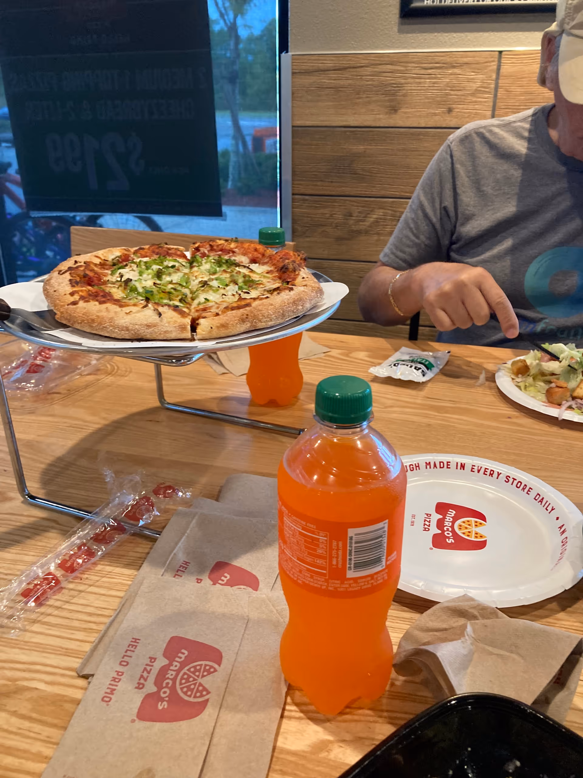 Pizza on a stand sits at a wooden dining table, with a masked person nearby.