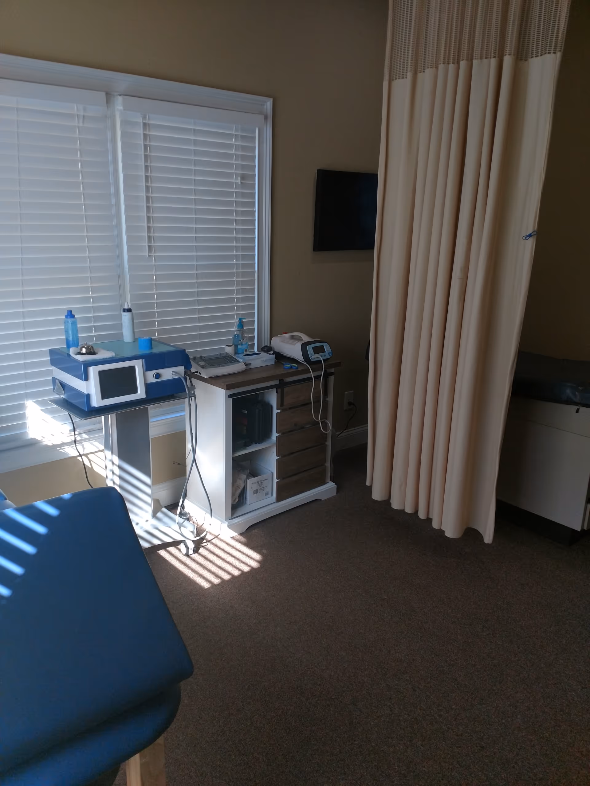 Clinical treatment area in a senior living facility with medical equipment, blinds, and a privacy curtain.