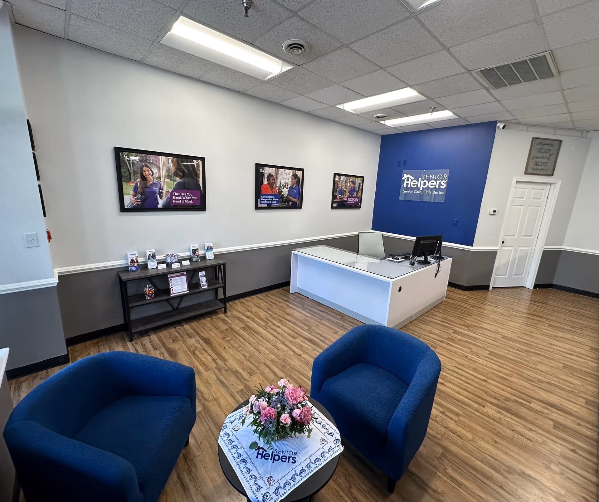 Senior Helpers reception area with blue wall, desk, framed photos, and blue chairs.