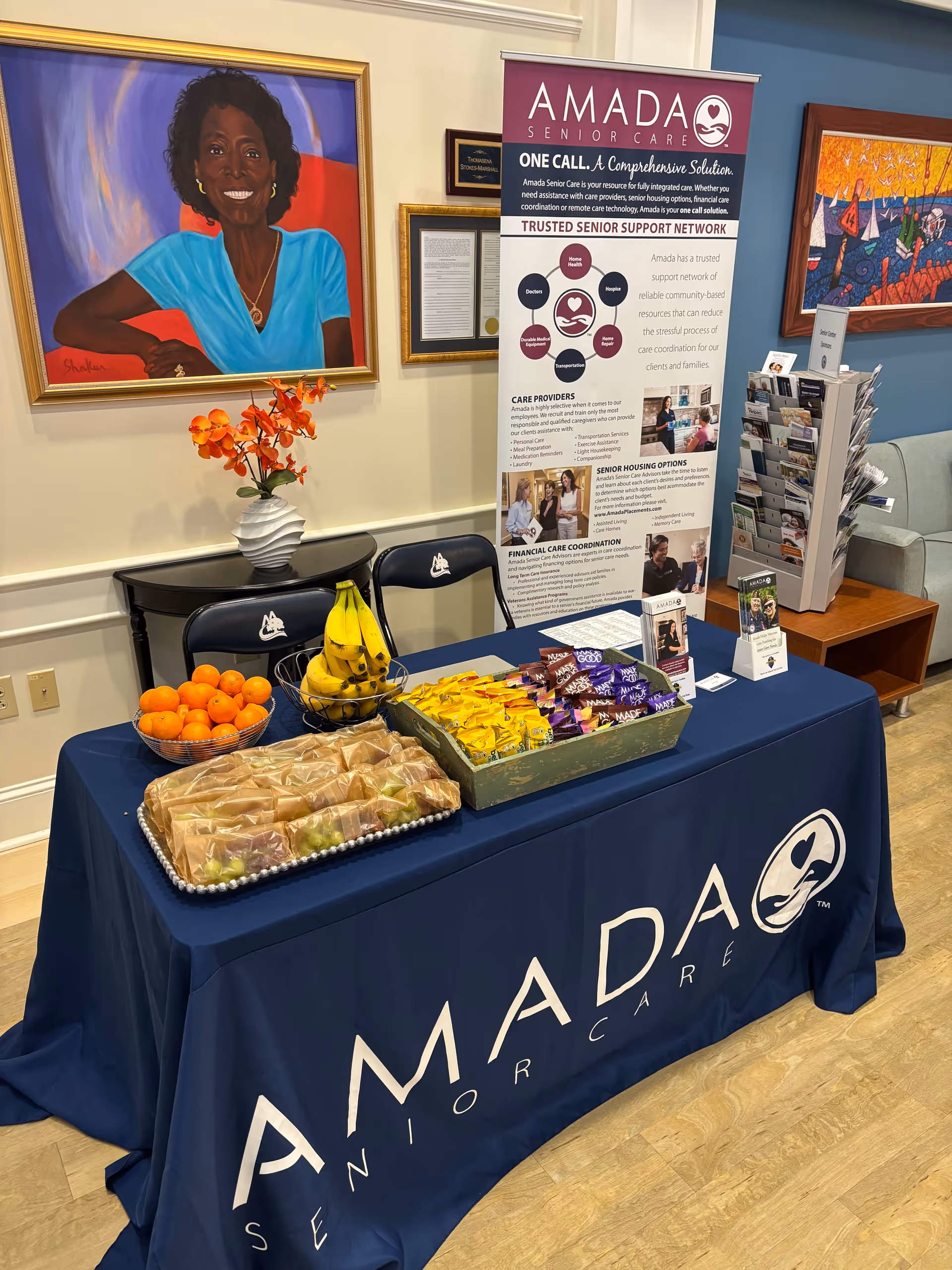 Amada Senior Care promotional table in a lobby with fruit, snacks, and brochures.