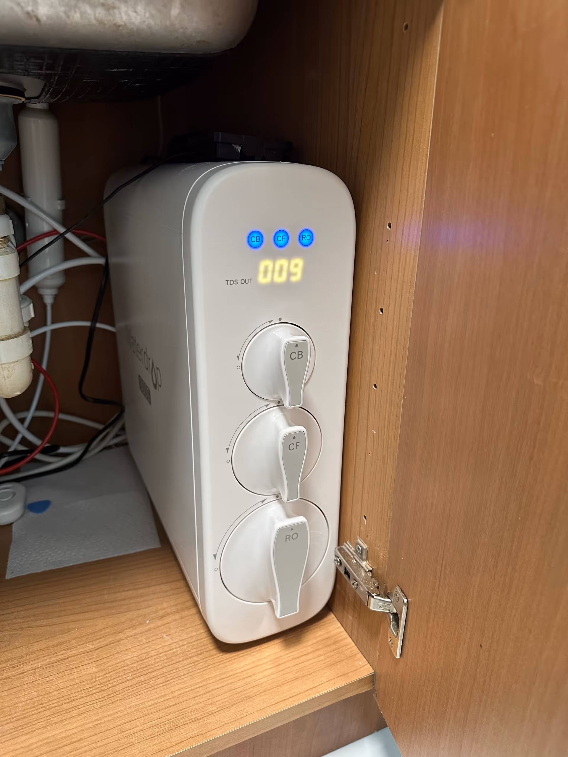 Under-sink water filtration device with three dial controls inside a wooden cabinet.
