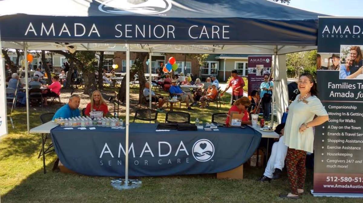 An Amada Senior Care outdoor booth under a tent with people seated at tables.