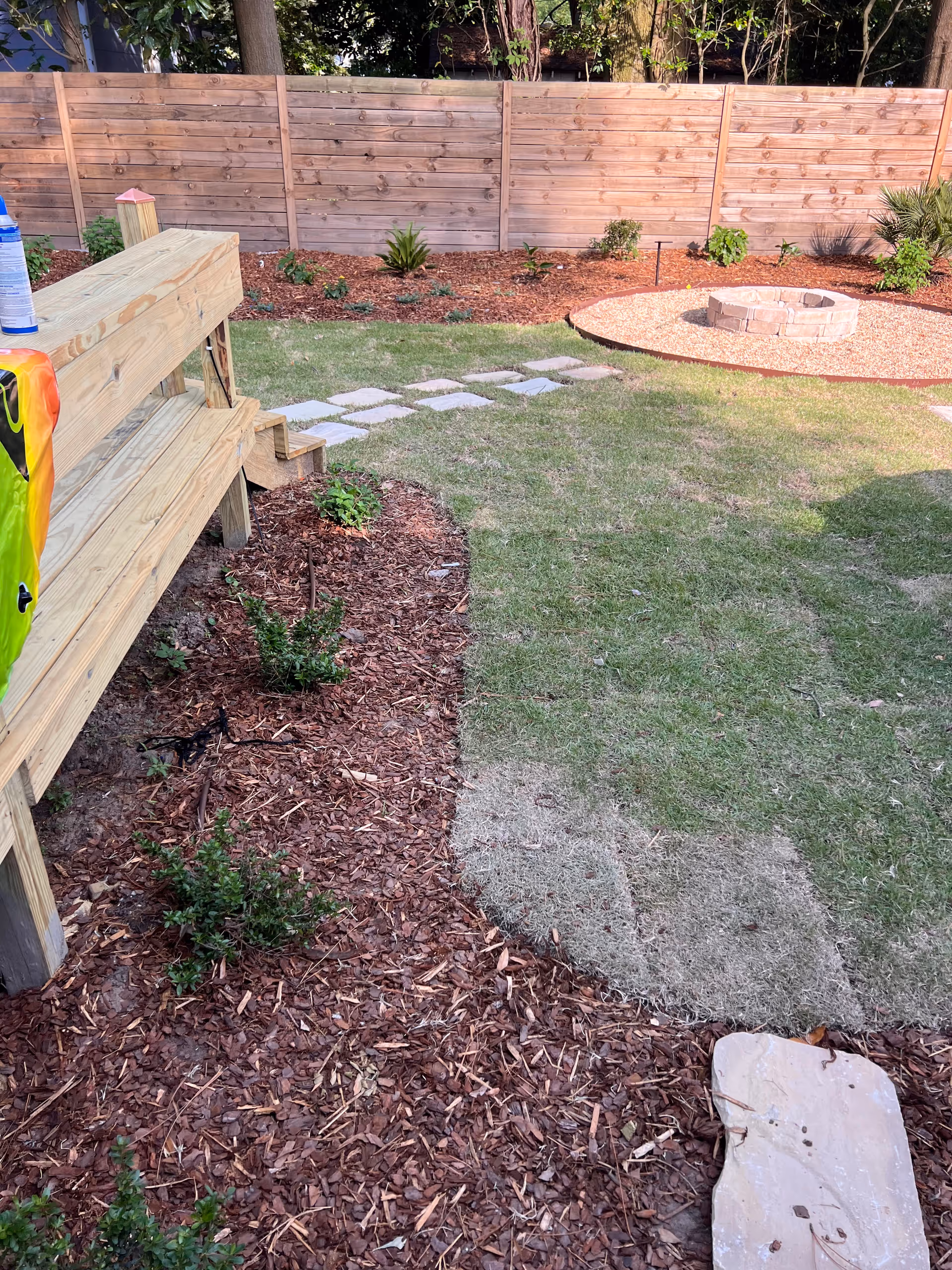 Backyard area with a wooden deck, stepping stones, mulch beds, and a circular fire pit.