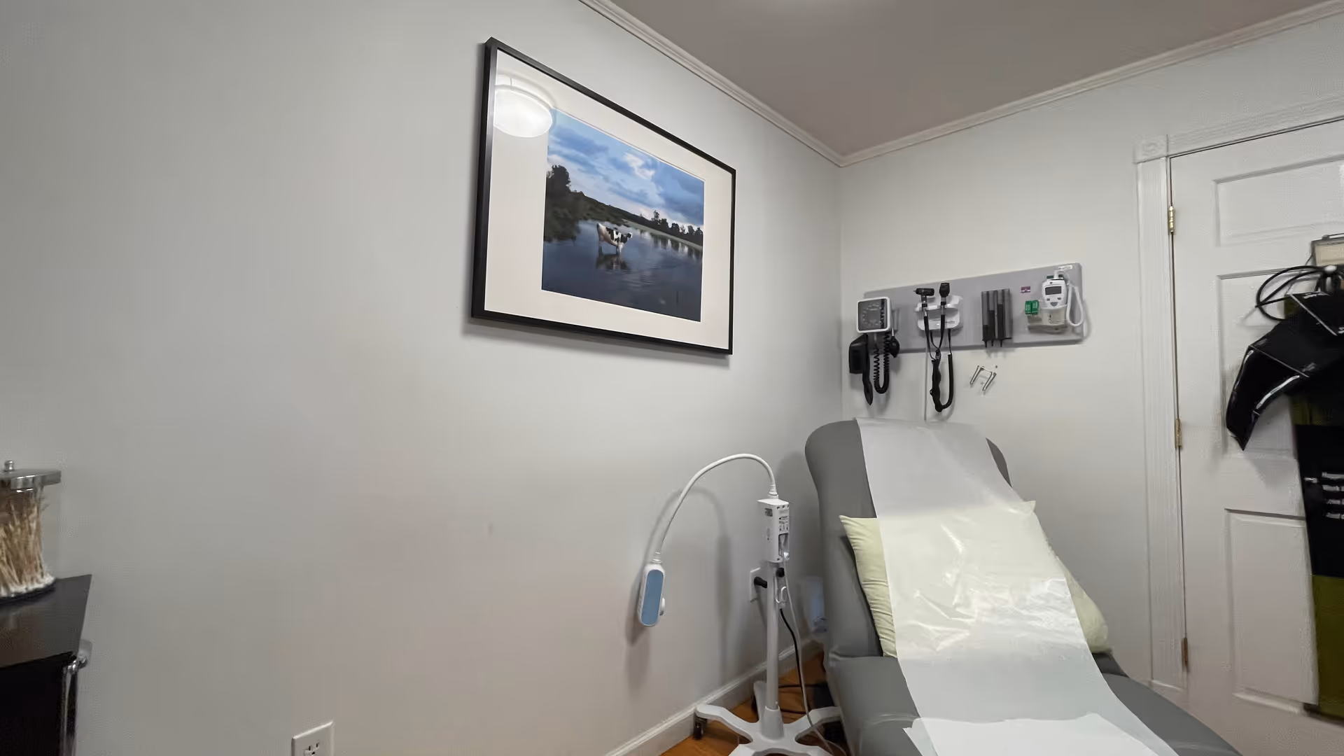 Medical examination chair in a small clinic room with wall-mounted equipment and framed art.