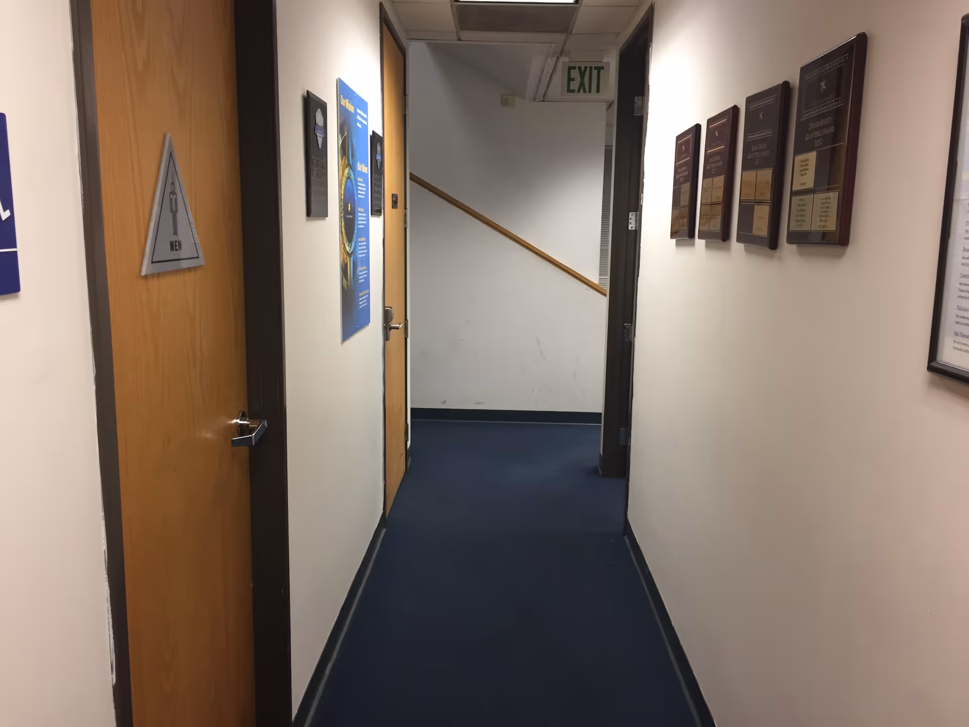 Narrow interior hallway in a facility with doors, wall plaques, and an exit sign.