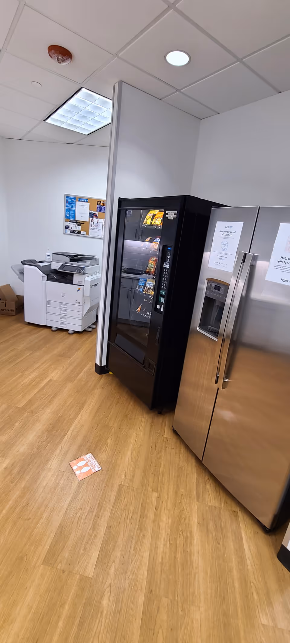 Vending machine, a stainless refrigerator, and a copier in a small staff break room.