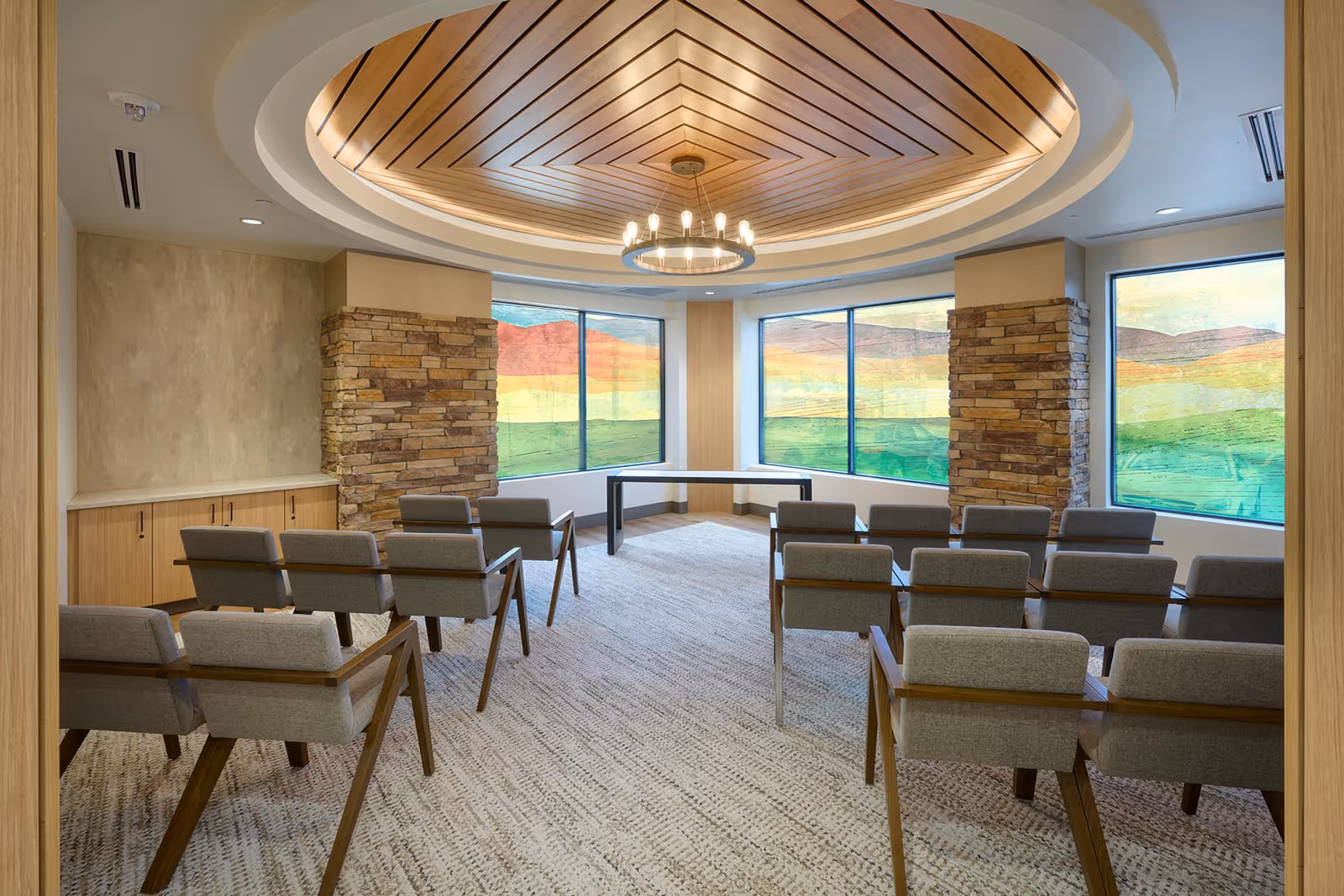 Sunlit conference room with circular wood-panel ceiling, chandelier, and rows of grey chairs.