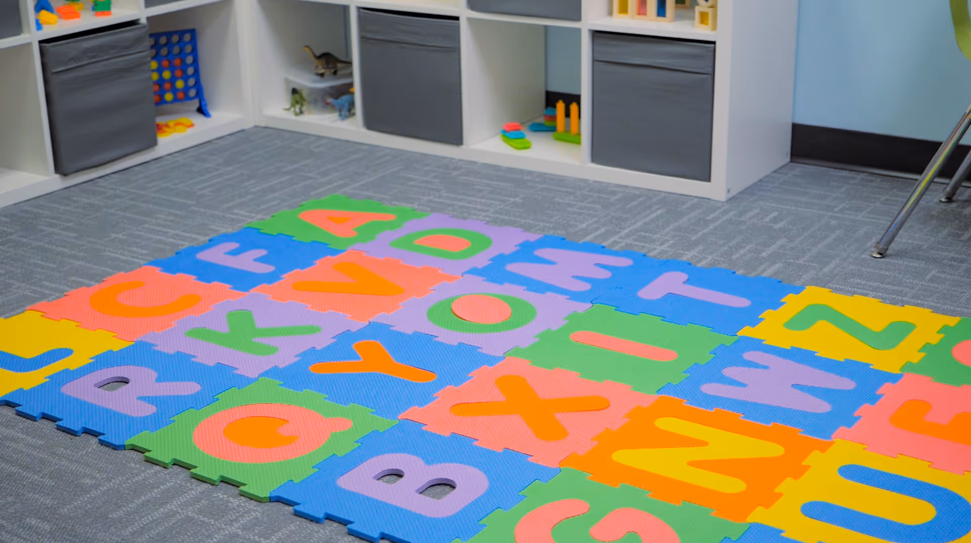 Colorful alphabet foam mats cover a bright play area with shelves along the wall.