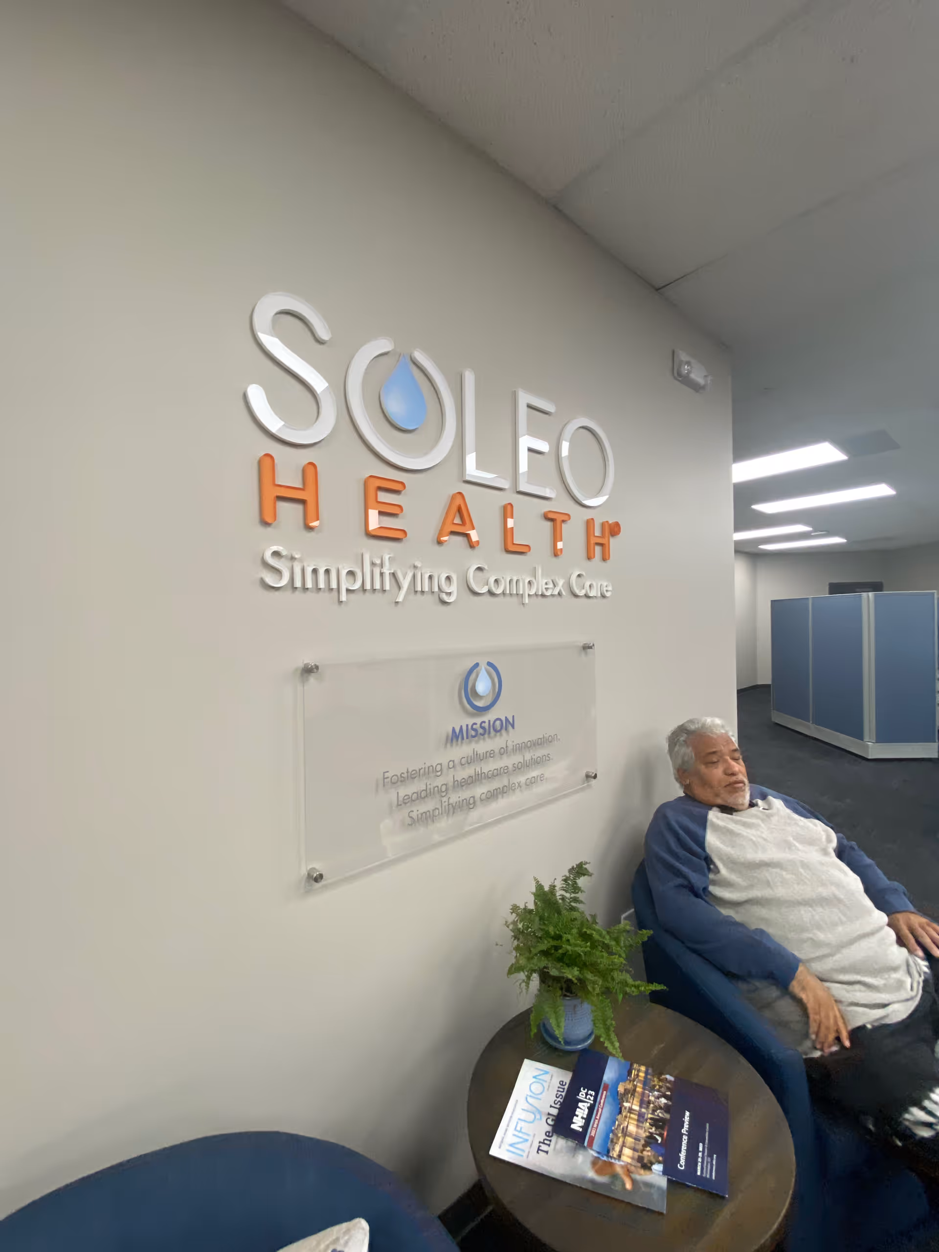 Lobby area with Soleo Health sign, a man seated in a blue chair beside a table.
