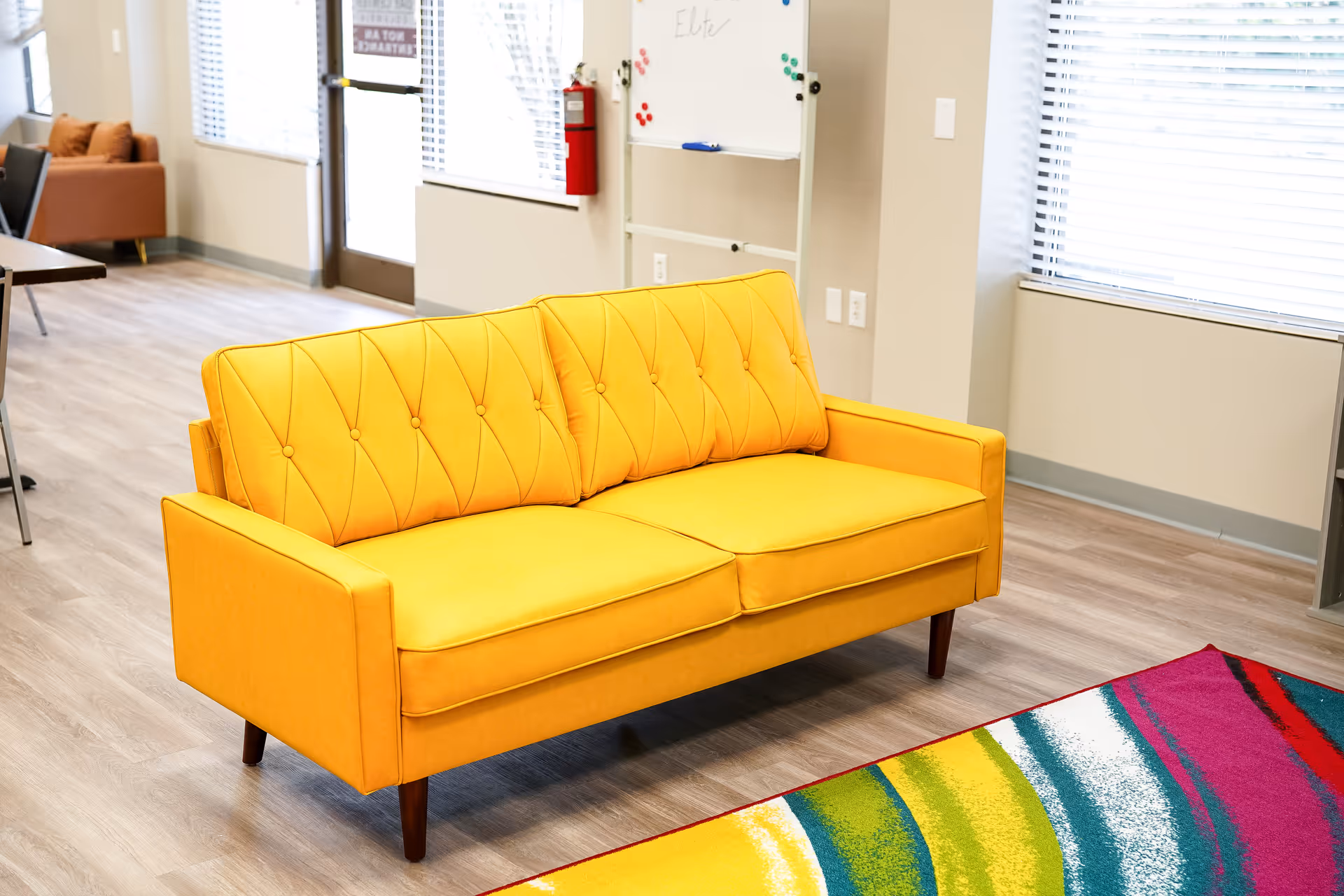 Bright yellow couch in a modern common area with windows and a colorful rug.