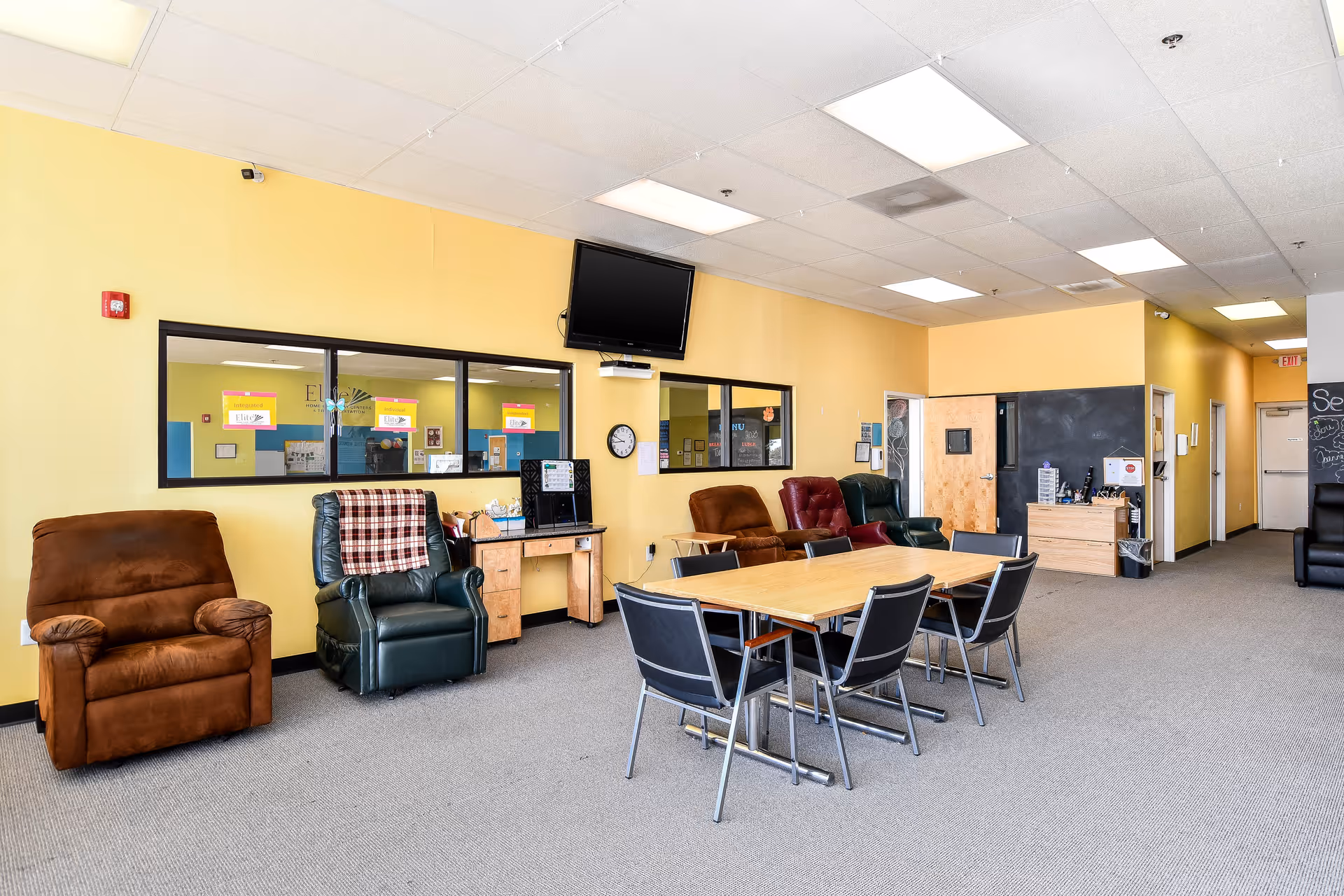 Bright yellow senior living lounge with recliners, a central table, and a wall-mounted TV.
