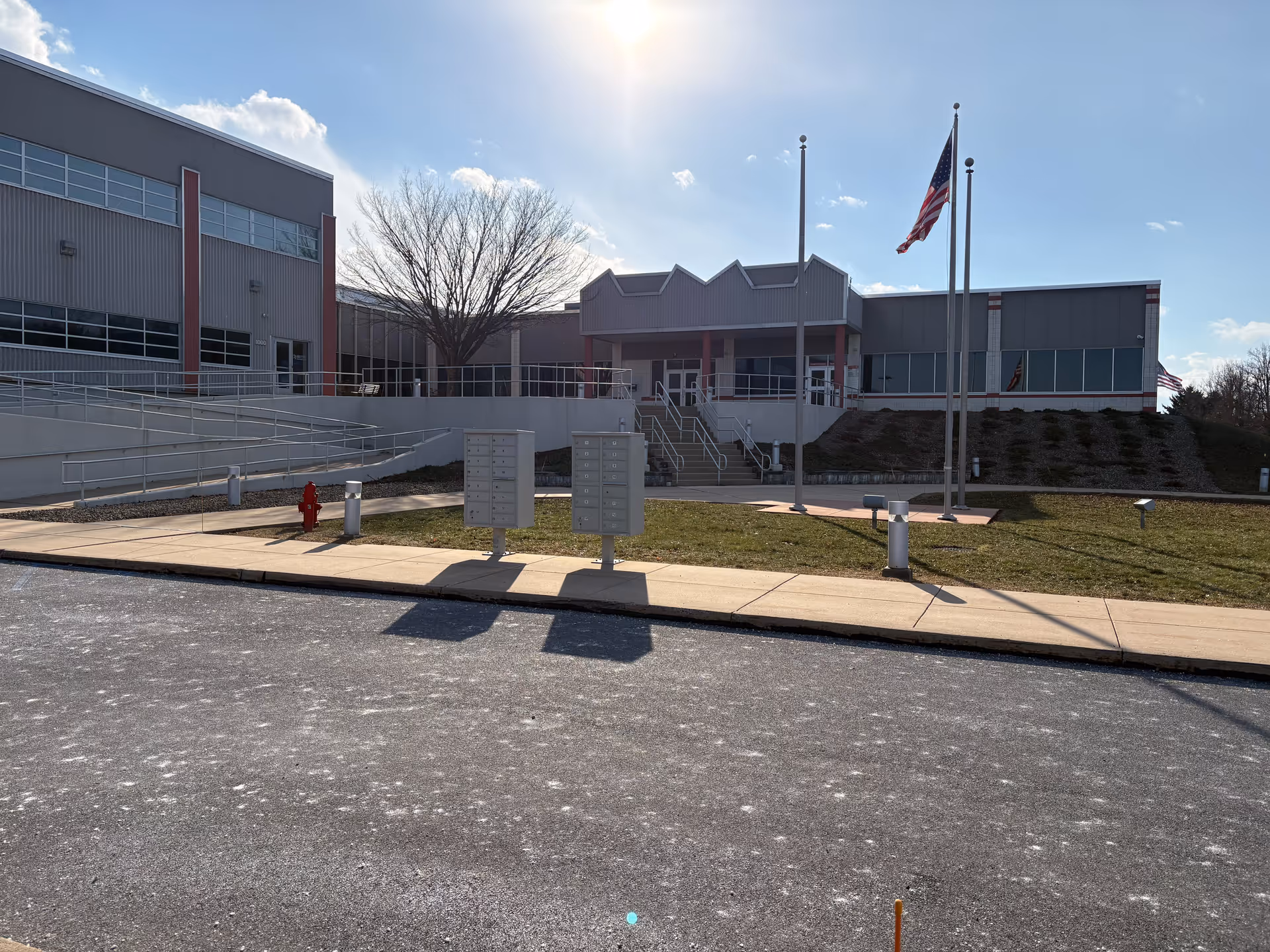 Modern facility exterior with entrance, stairs, flagpoles, and a bright, clear sky.