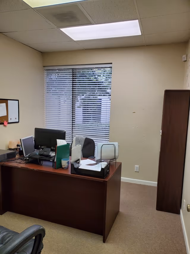 Office workspace with desk, computer, filing trays, and tall cabinet beside a window.