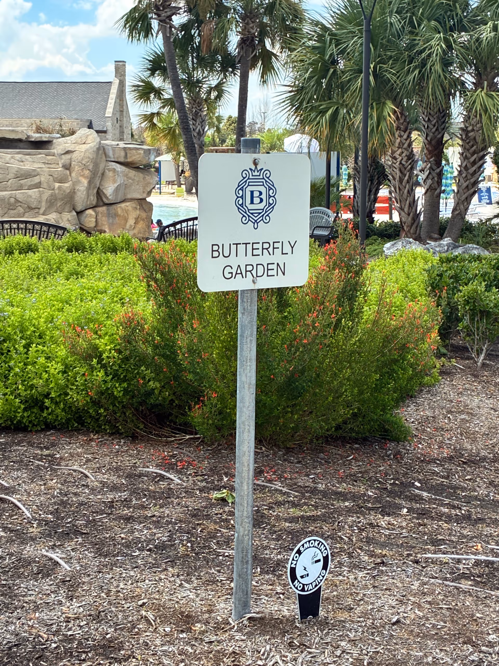 Outdoor garden area with Butterfly Garden sign, palm trees, rocks, and a no-smoking marker.