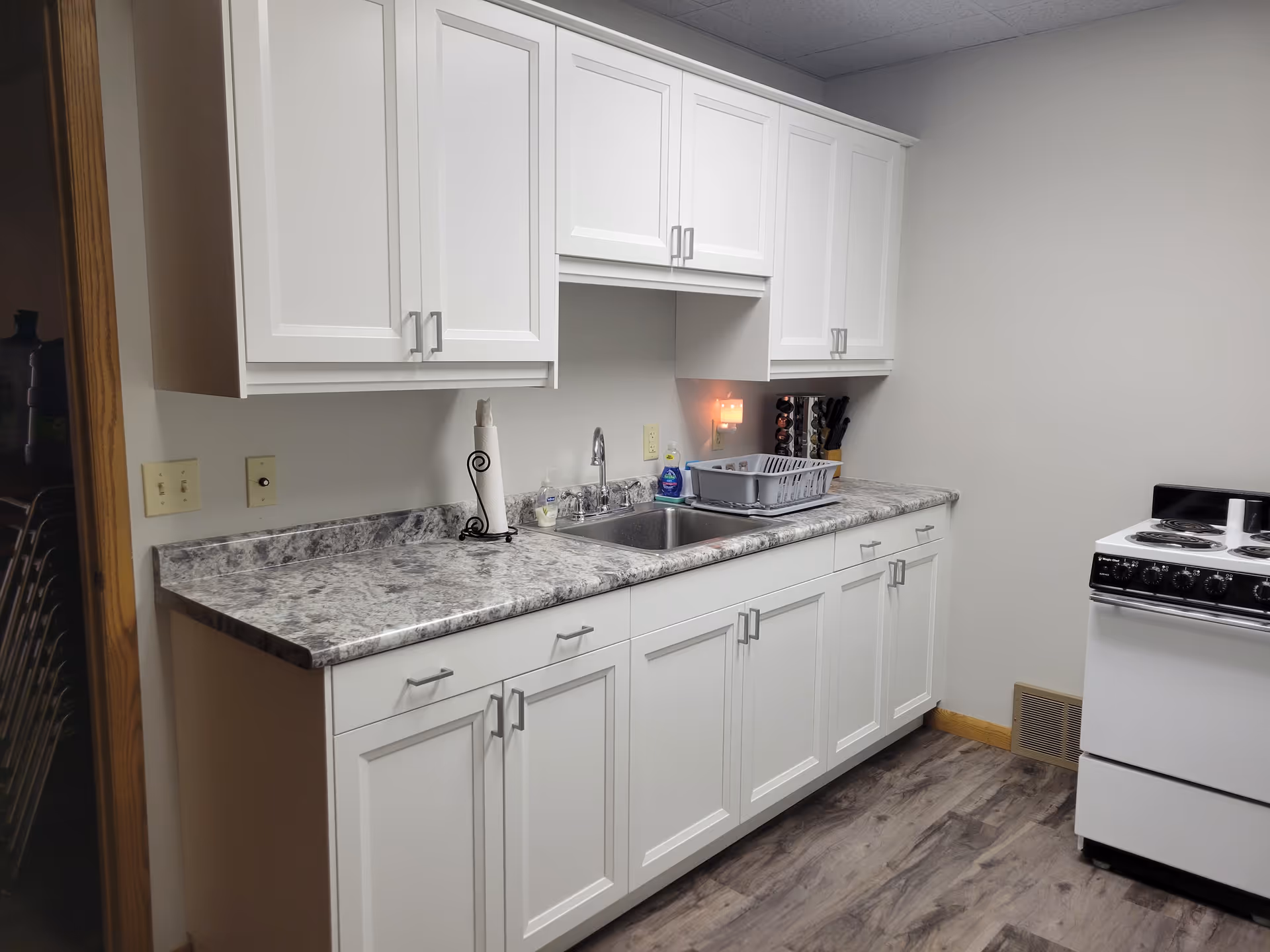 White kitchen with cabinets, sink, dish rack, and stove along marble countertop.