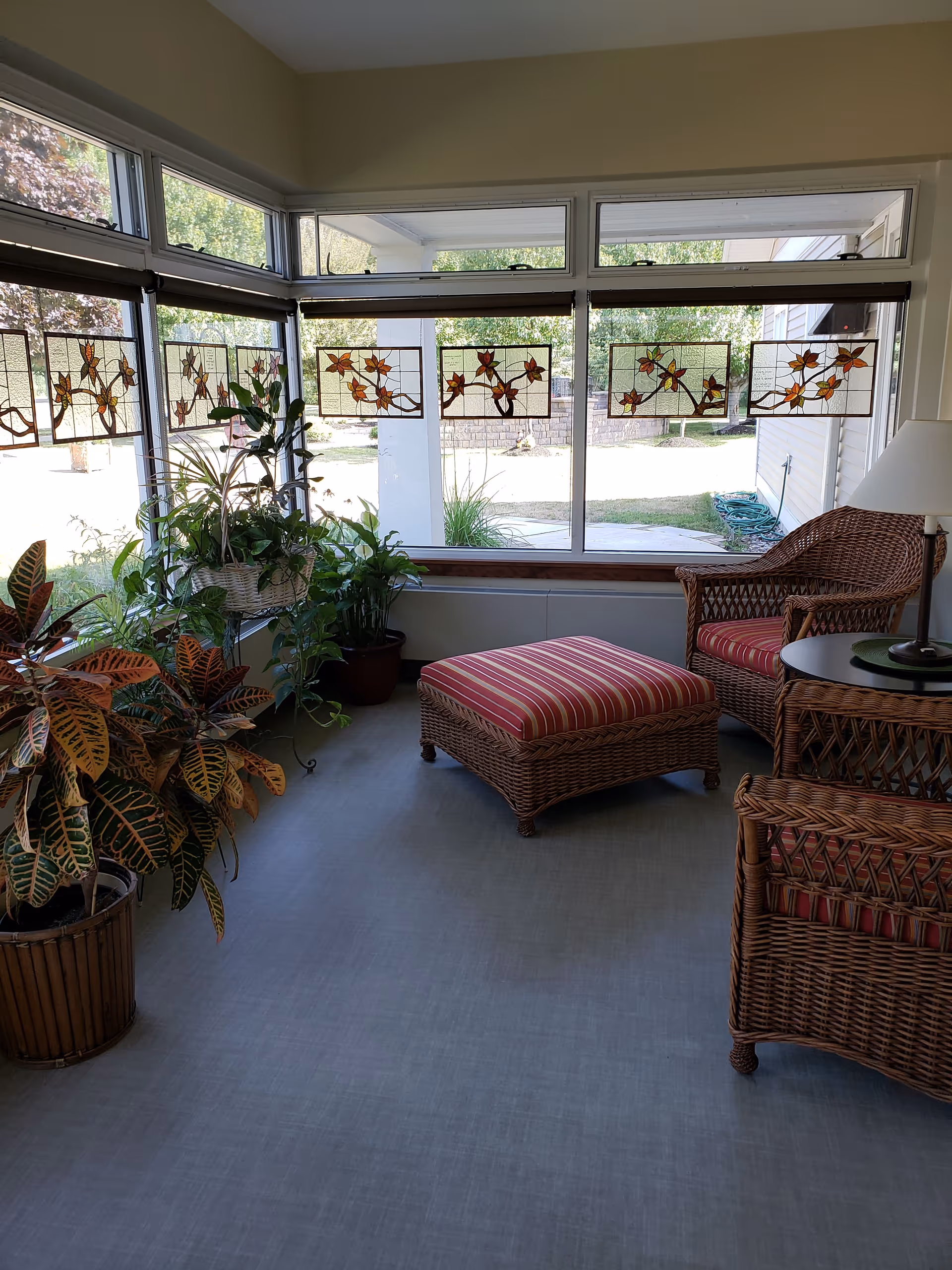 Bright sunroom lounge with wicker chairs, ottoman, and potted plants by large windows.