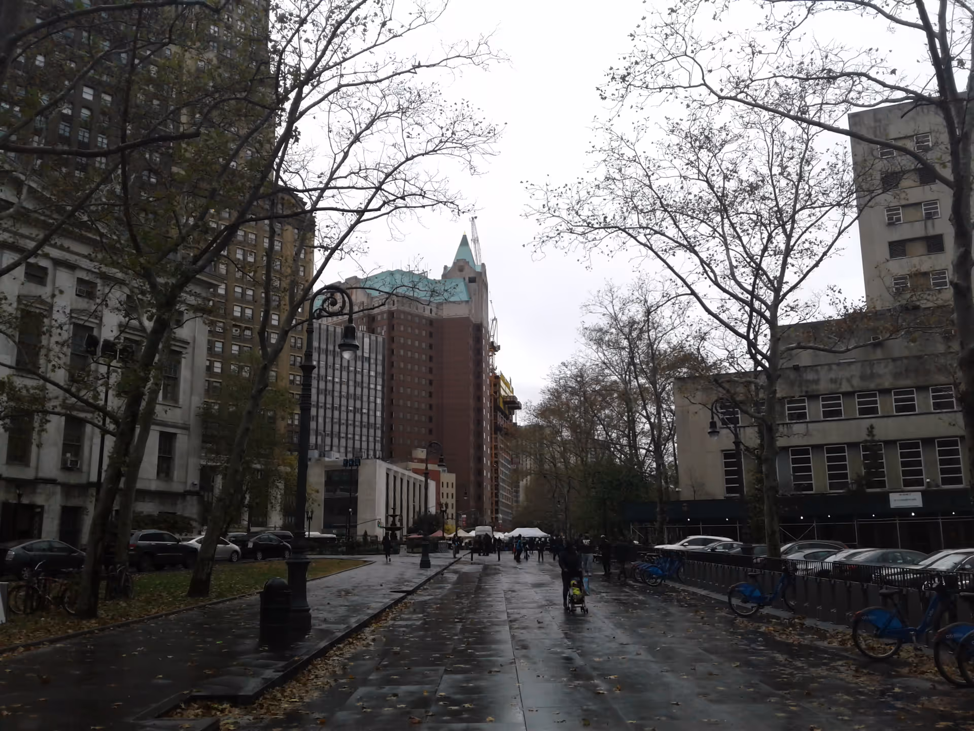 Rainy urban street with bare trees, tall buildings, and pedestrians on a wet sidewalk.