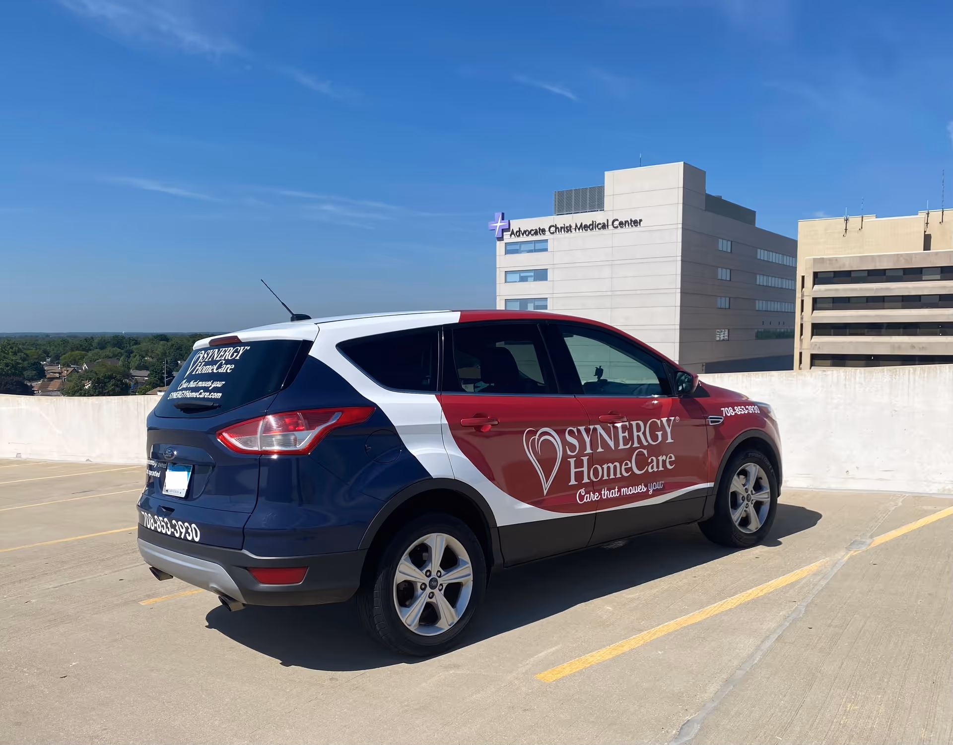 A Synergy HomeCare van parked on a rooftop parking lot with a hospital building in background.