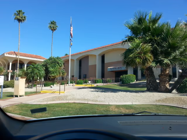 Exterior view of a beige senior care building with arches, palm trees, and a flagpole.