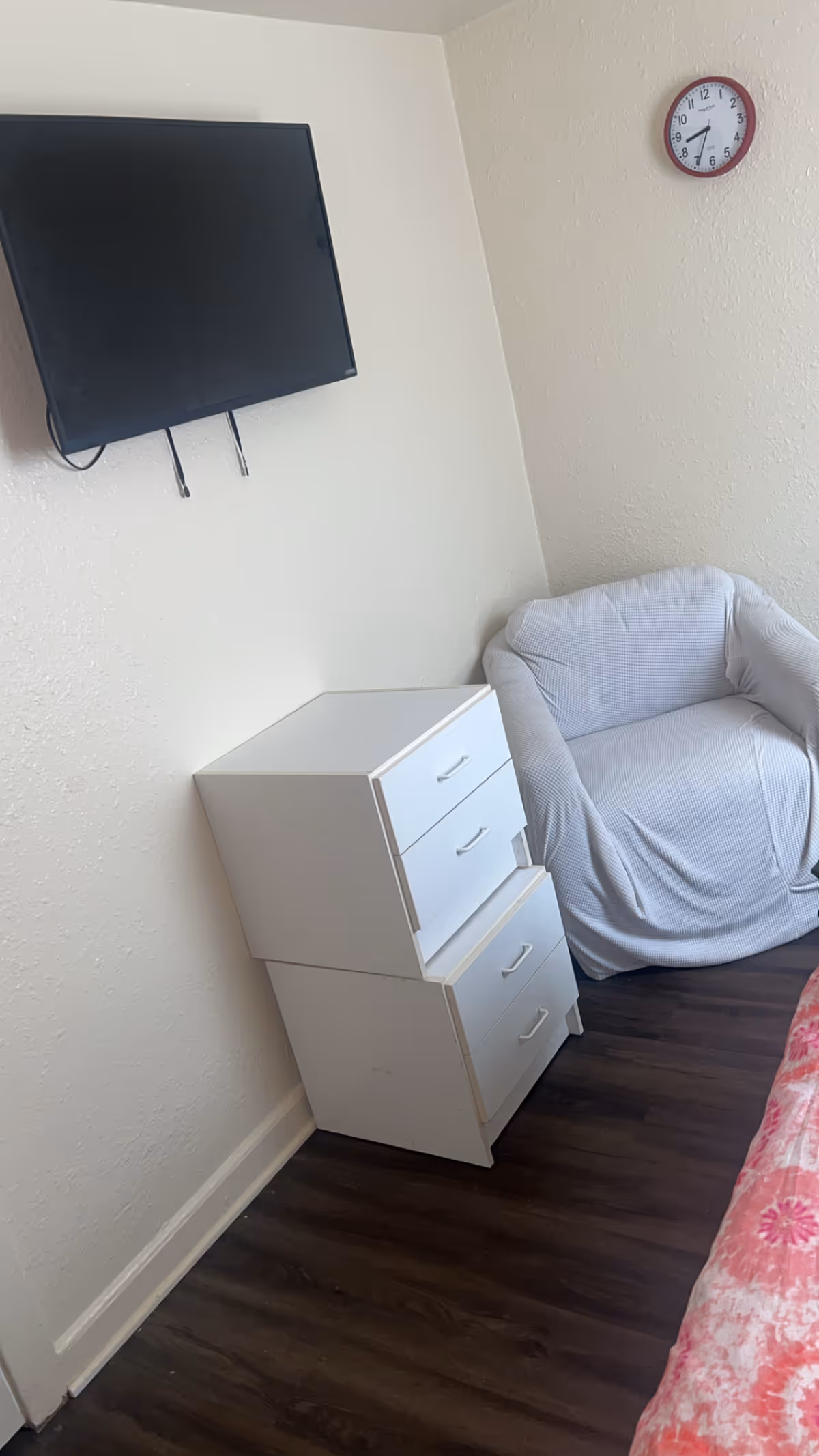 Corner bedroom with mounted TV, white dresser, gray cushioned chair, clock, and wood floor.