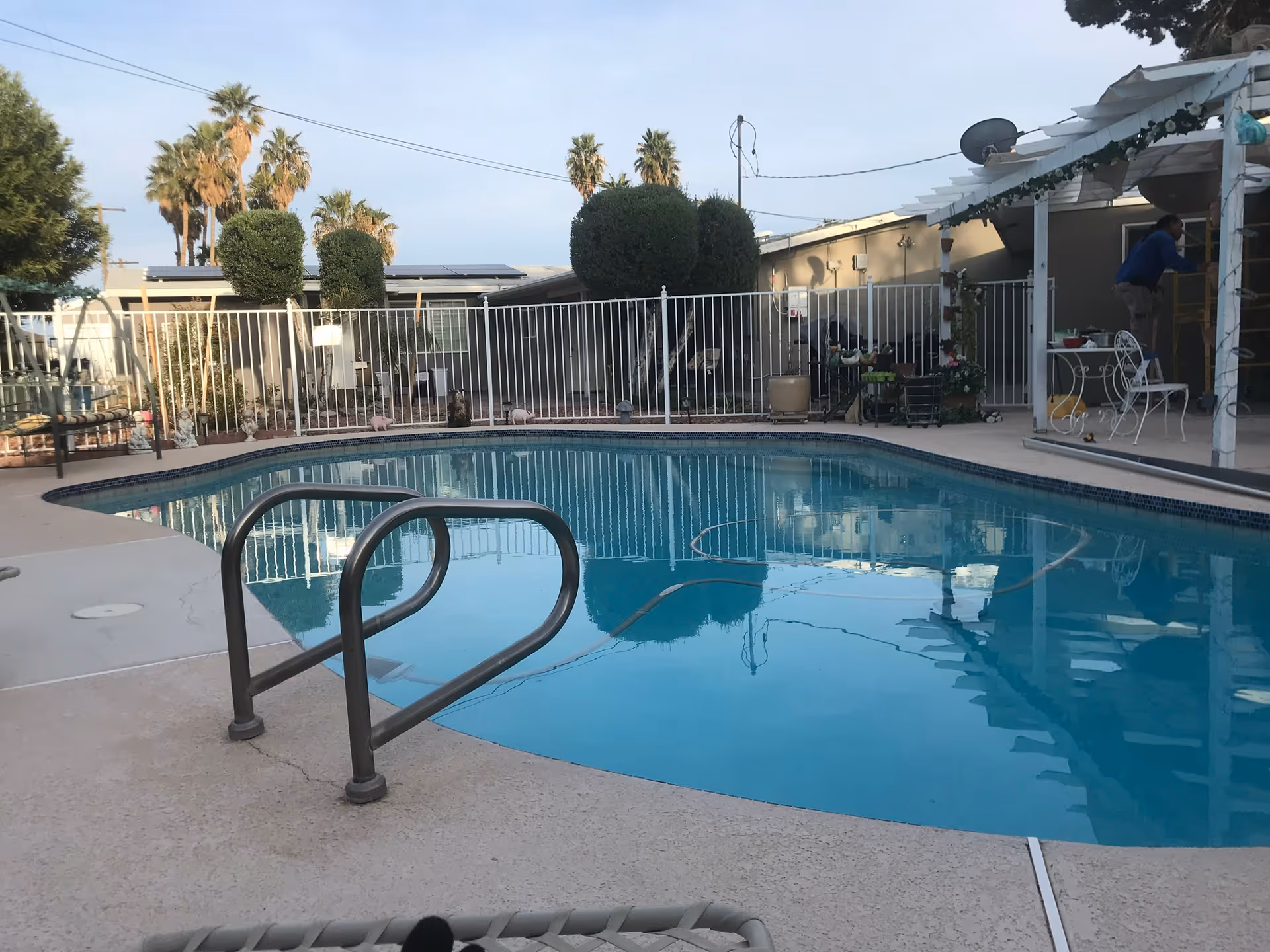 Outdoor swimming pool at a senior care facility with fencing, palm trees, and patio seating.