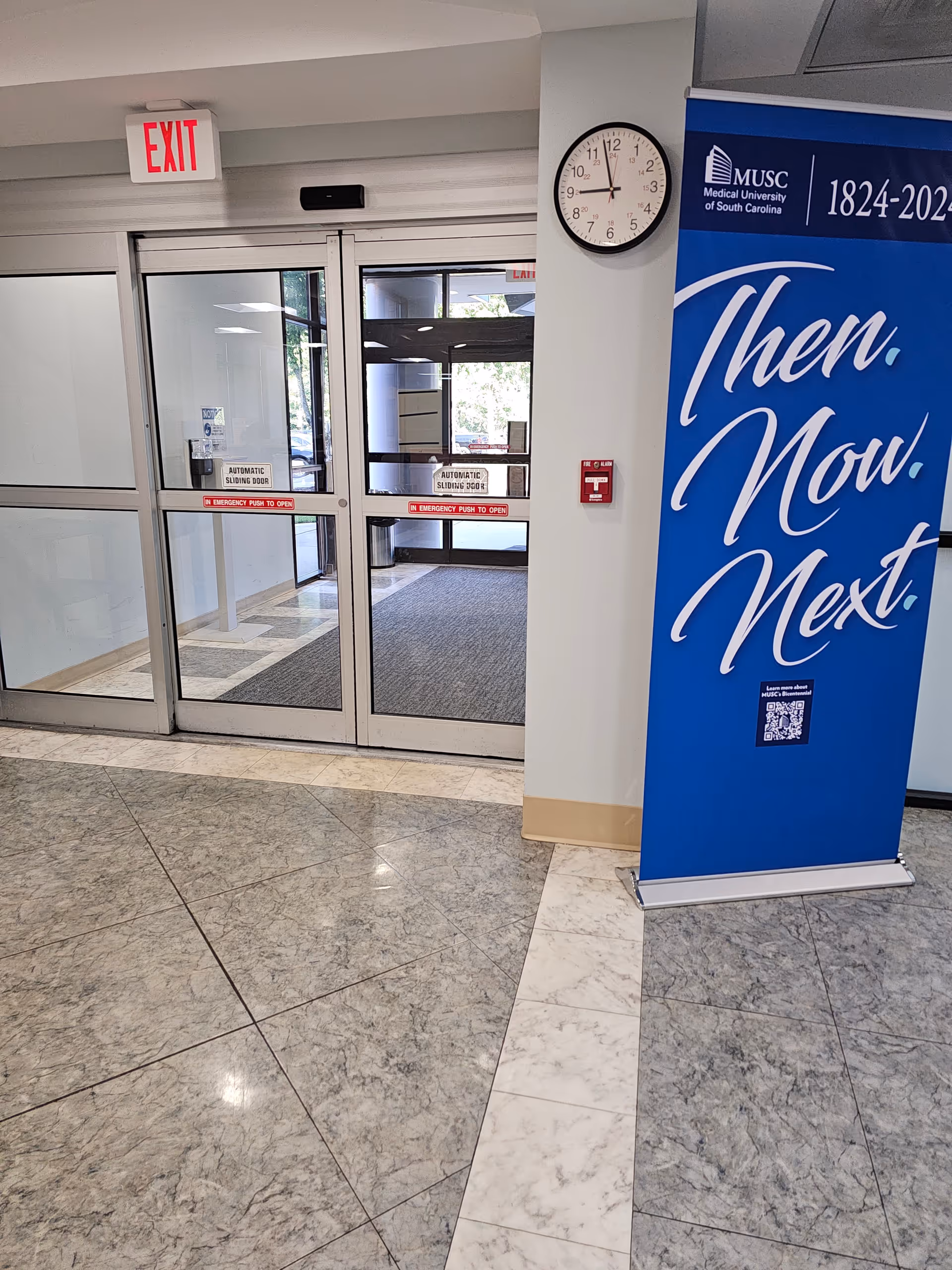 Interior hospital lobby with glass doors, exit sign, clock, and a blue banner