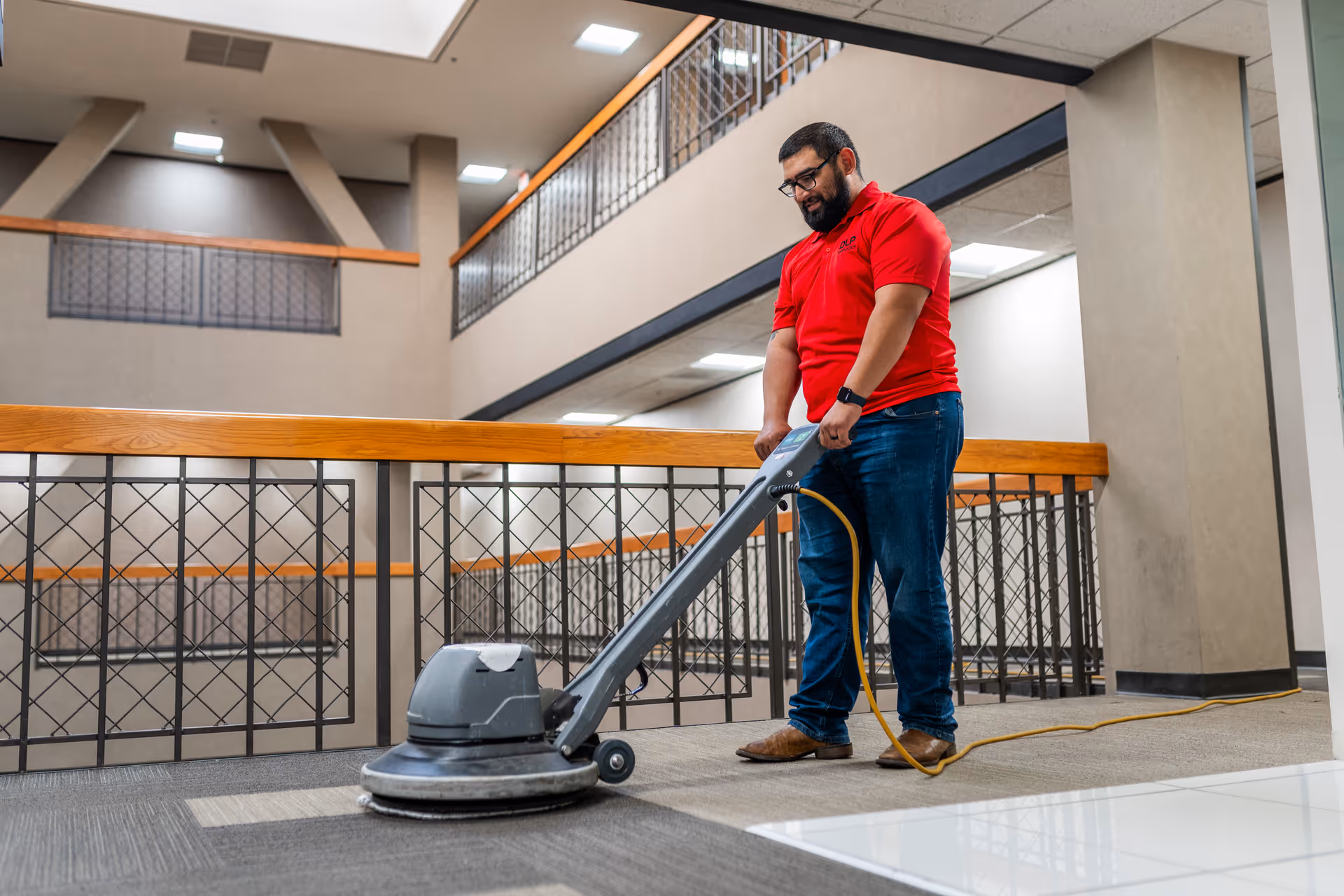 A man in a red shirt uses a floor polishing machine in a spacious indoor hallway.