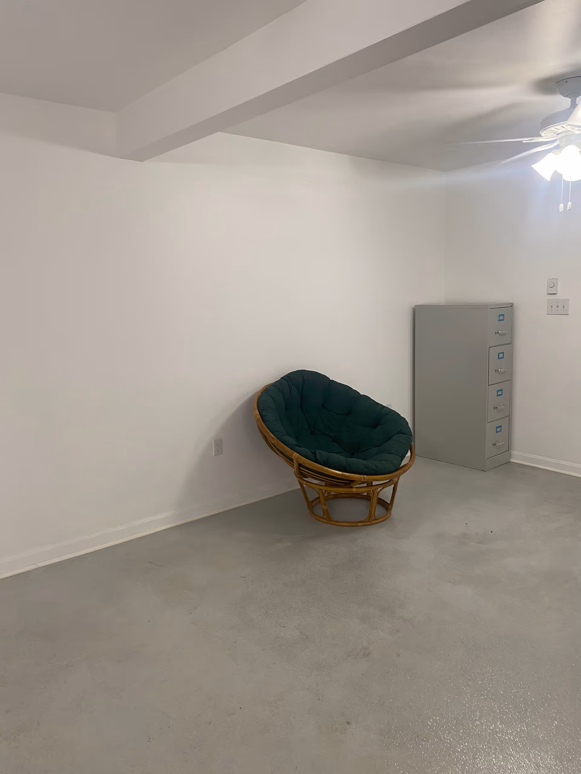 Bare white interior room with a green cushioned wicker chair and a gray filing cabinet.