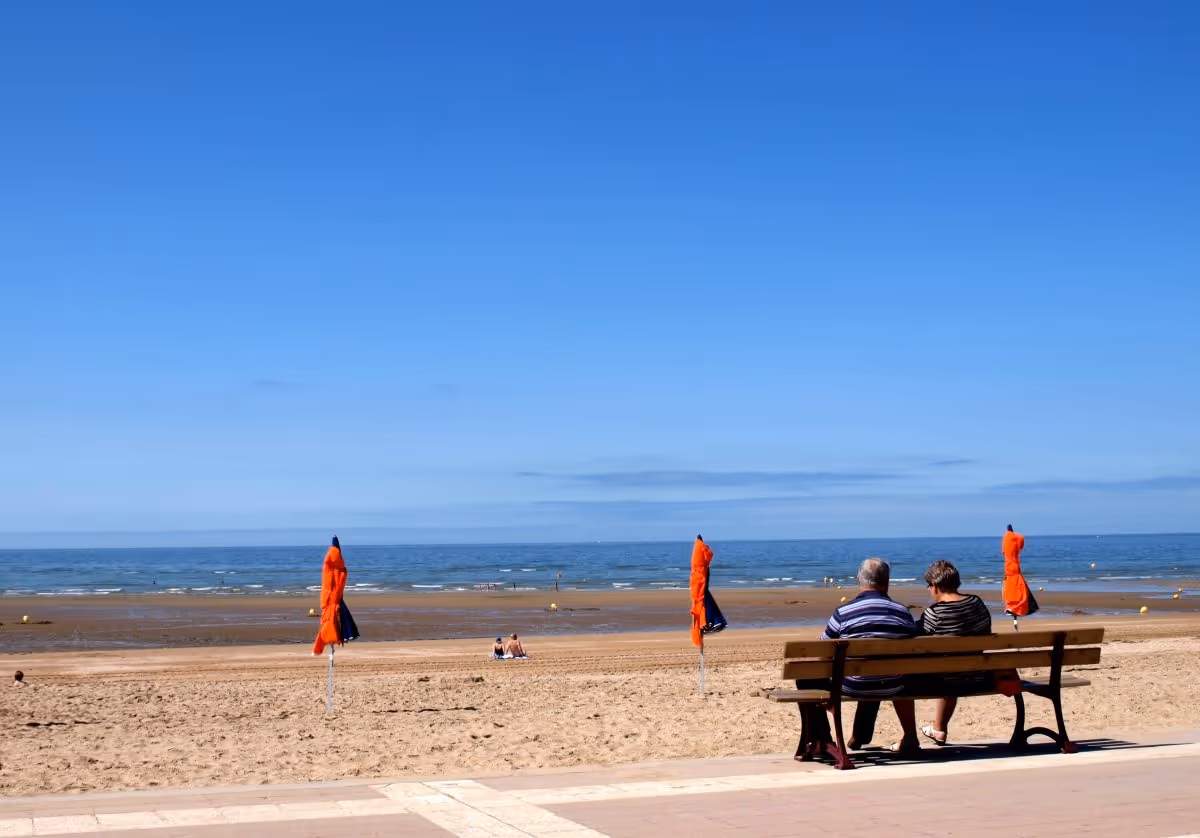 Two seniors sit on a bench by the beach with orange umbrellas and ocean view.