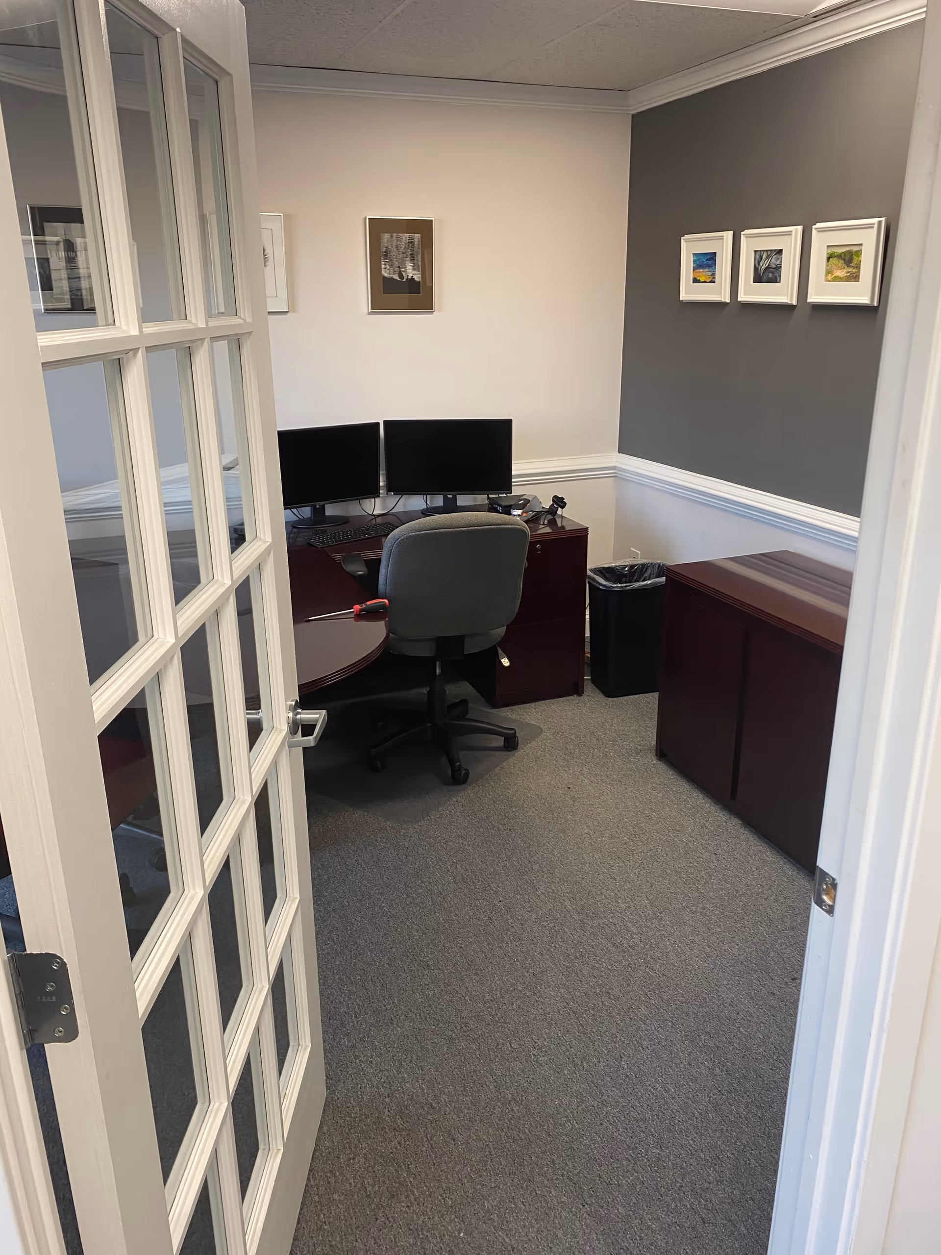 Small office with dual monitors, desk, chair, filing cabinet, and framed wall art.
