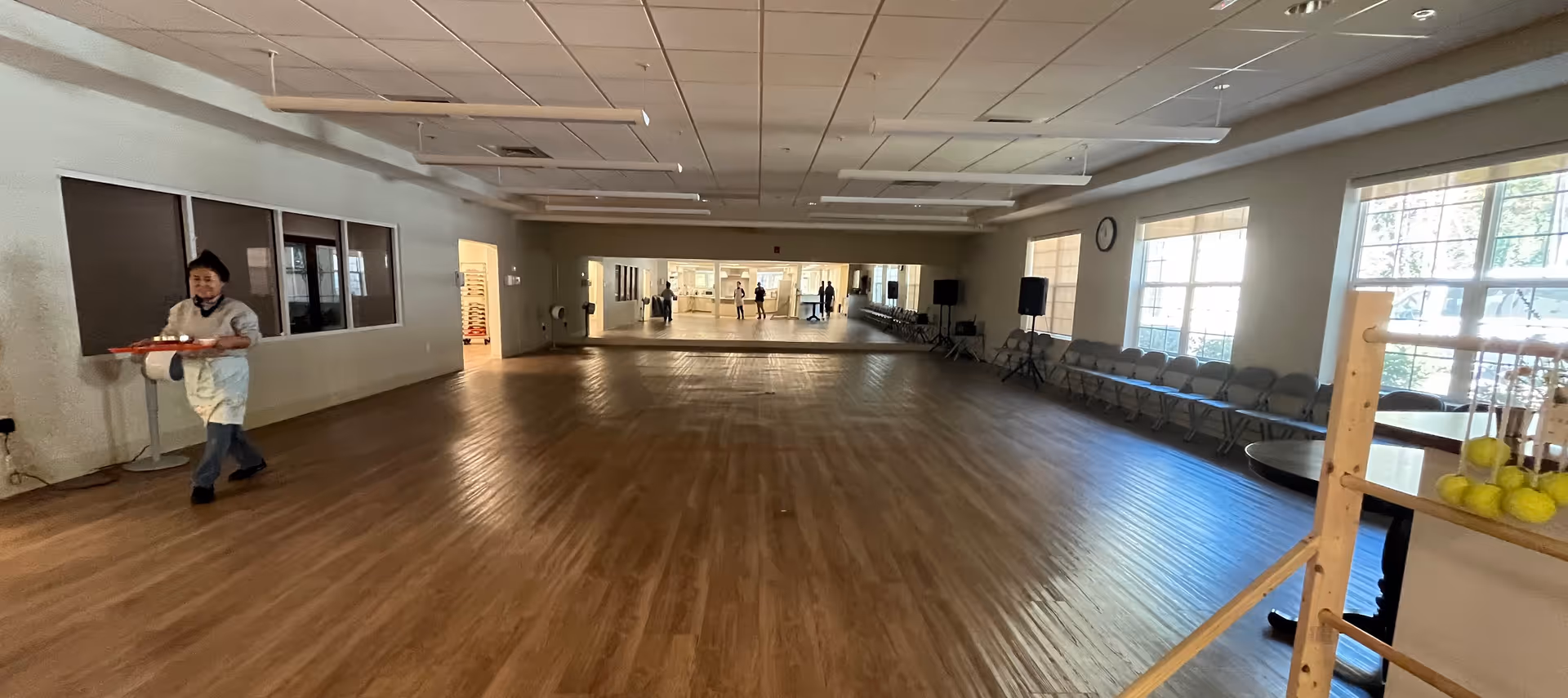A wide multipurpose room in a senior living facility with a staff member carrying a tray.