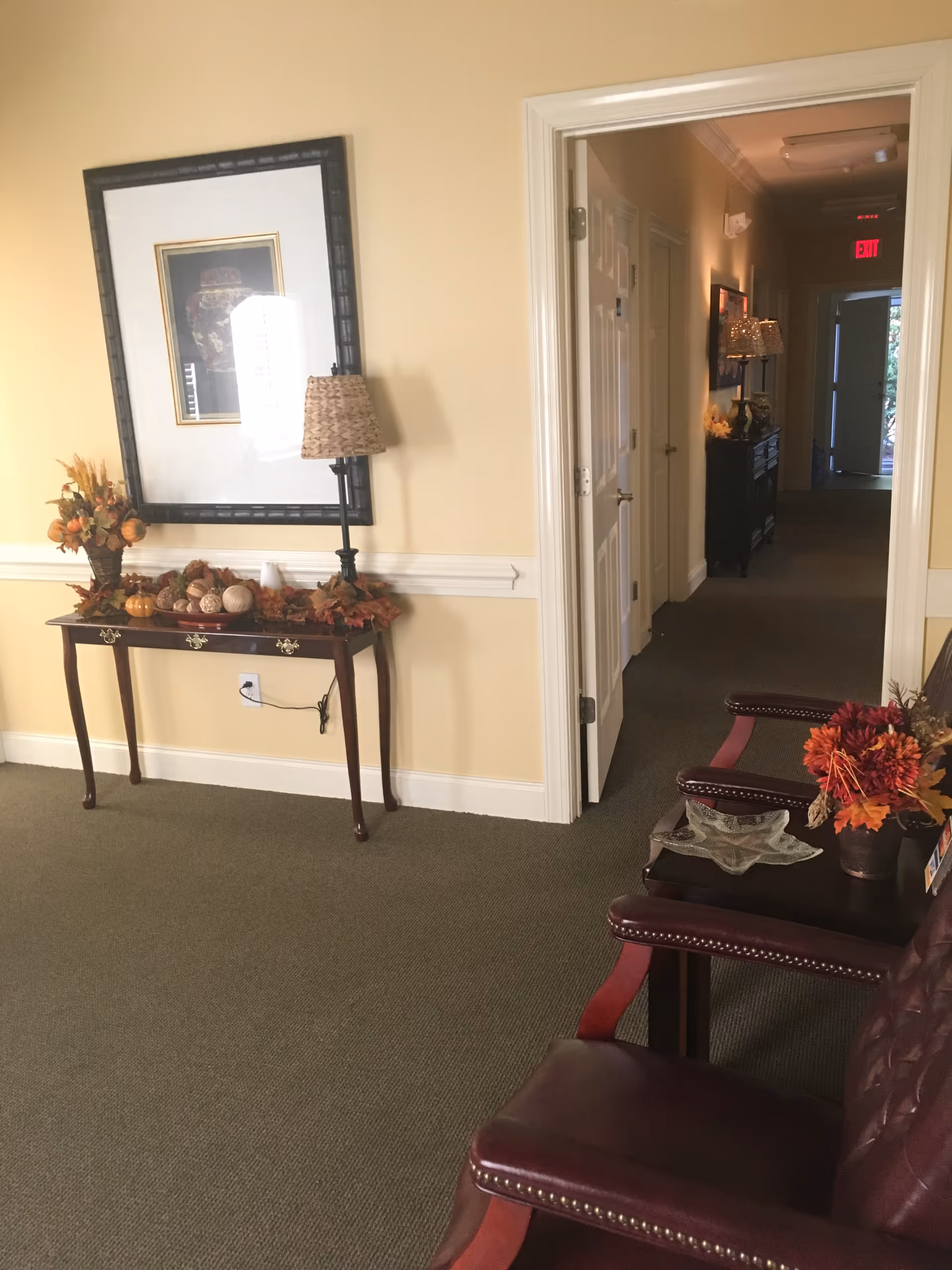 Interior hallway in a senior living facility with a console table, fall decor, and burgundy chairs.