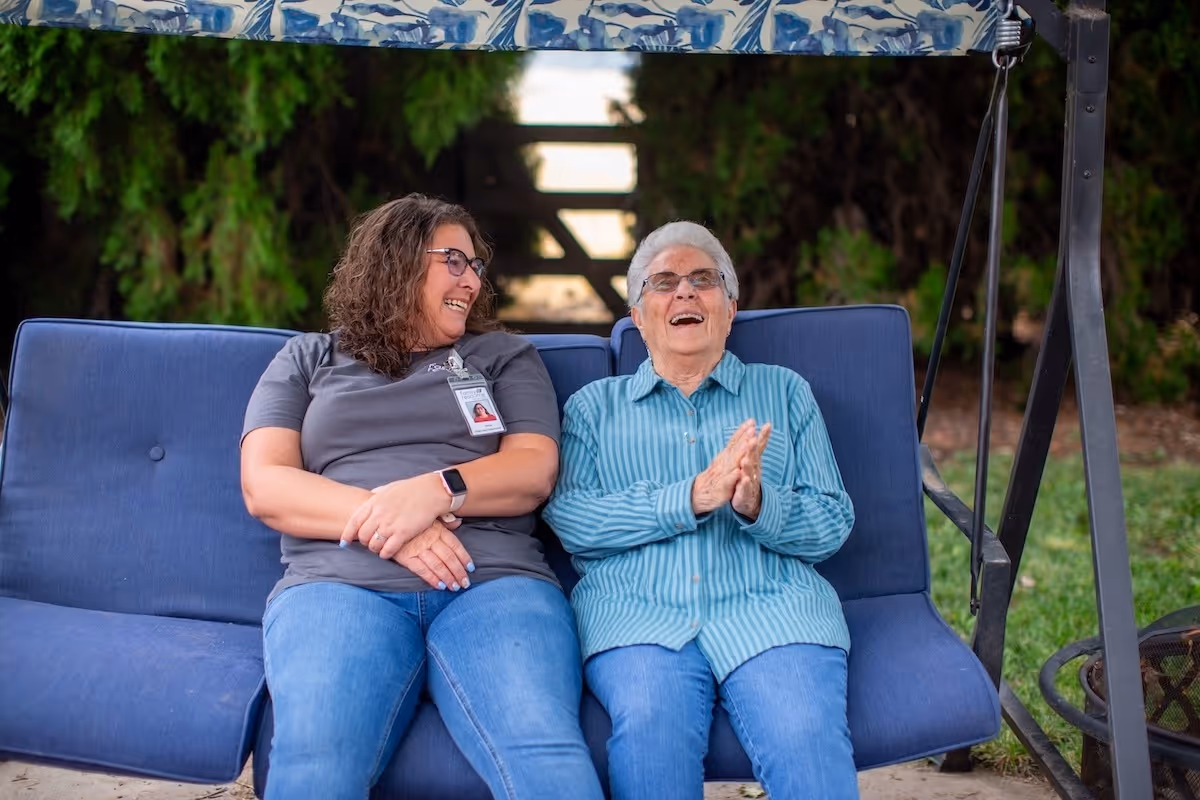 Two smiling women sit on a blue outdoor swing bench, chatting and enjoying time outside.