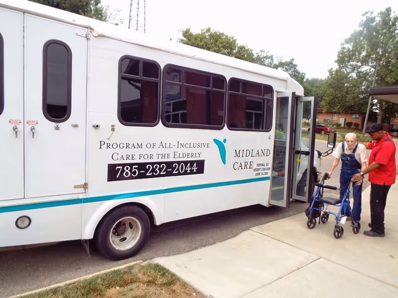 A Midland Care shuttle bus parked outside with elderly resident using a walker and a caregiver.