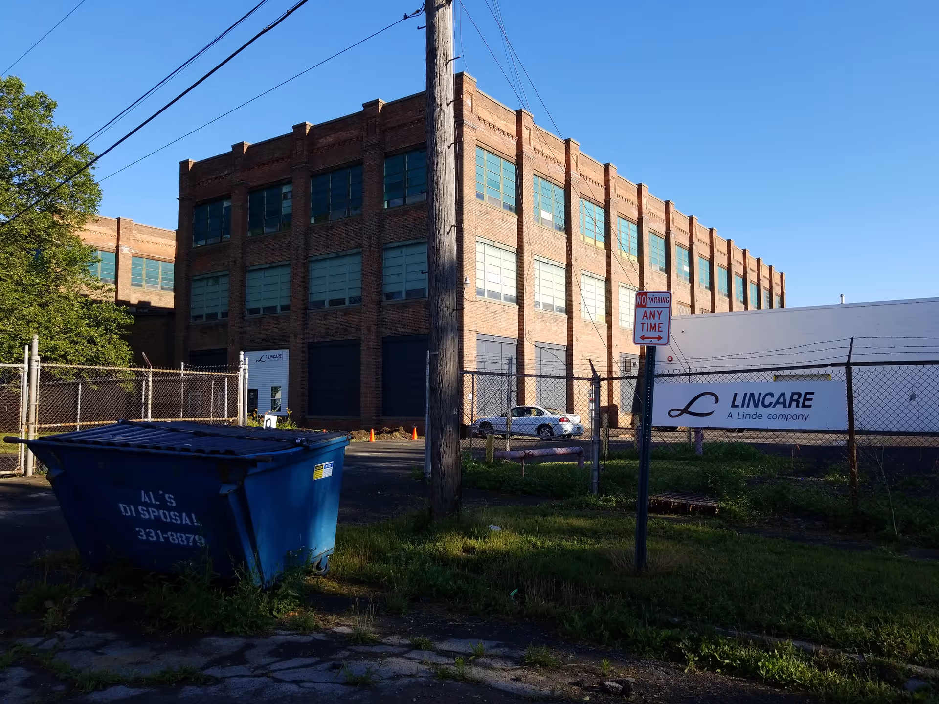 Industrial brick building with a LINCARE sign, chain-link fence, dumpster, and utility pole in foreground.