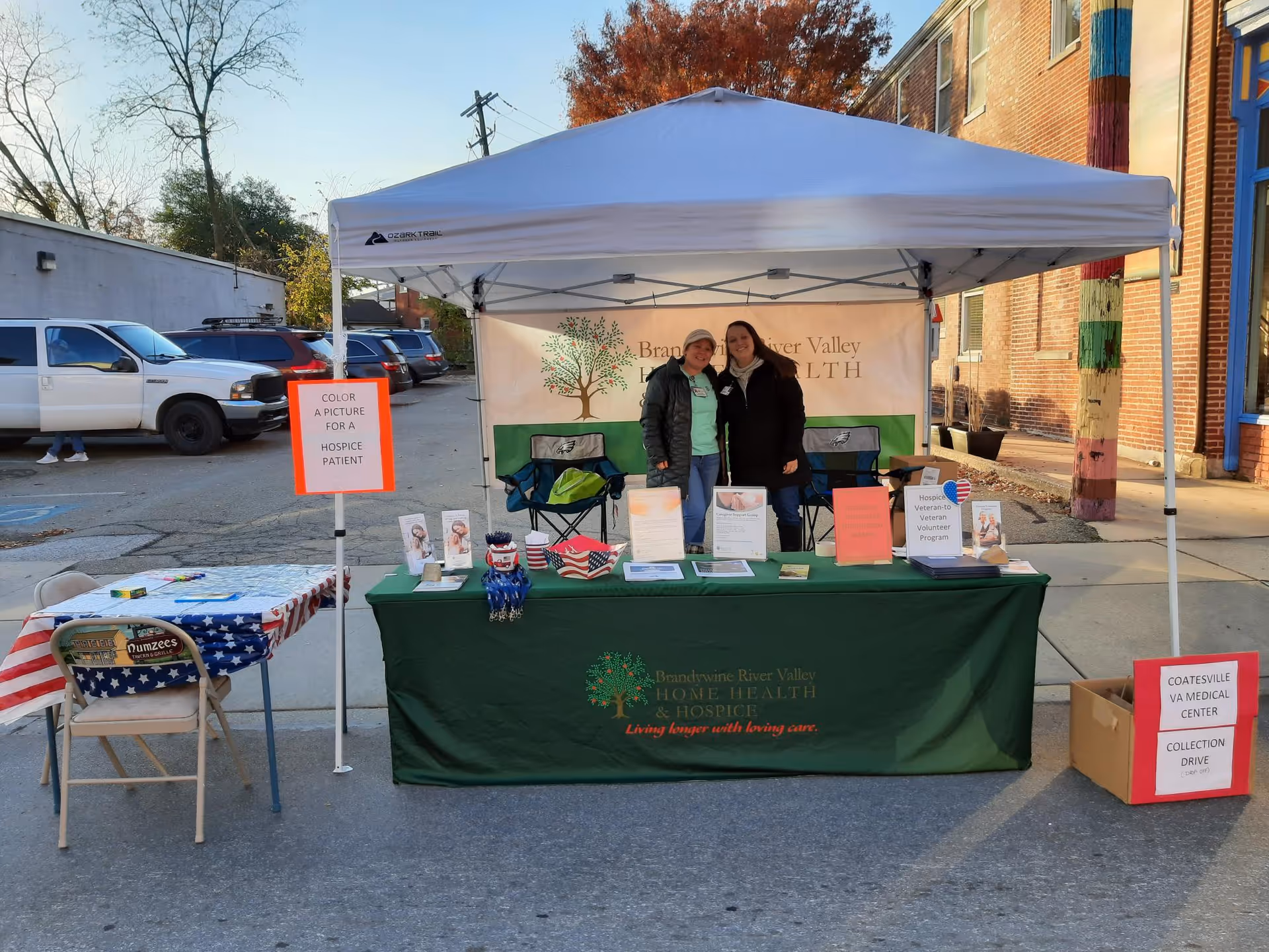 Outdoor booth with two staff under a white canopy promoting hospice services.