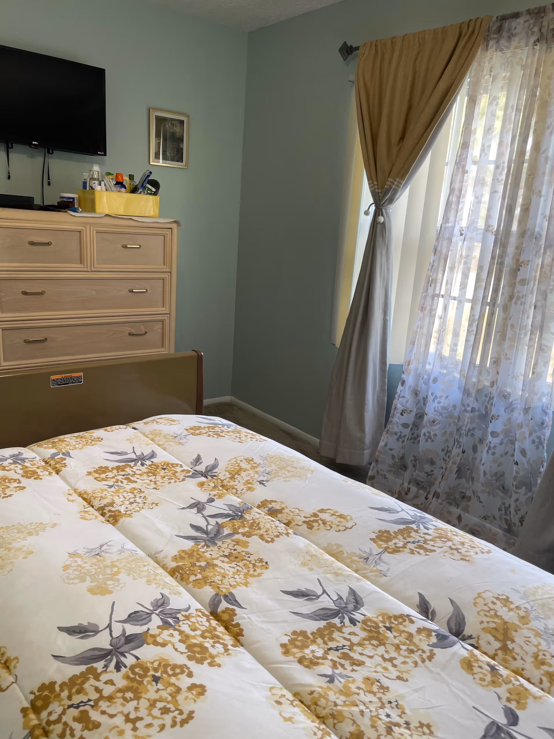 Senior living facility bedroom with floral bedspread, dresser, and wall-mounted TV.