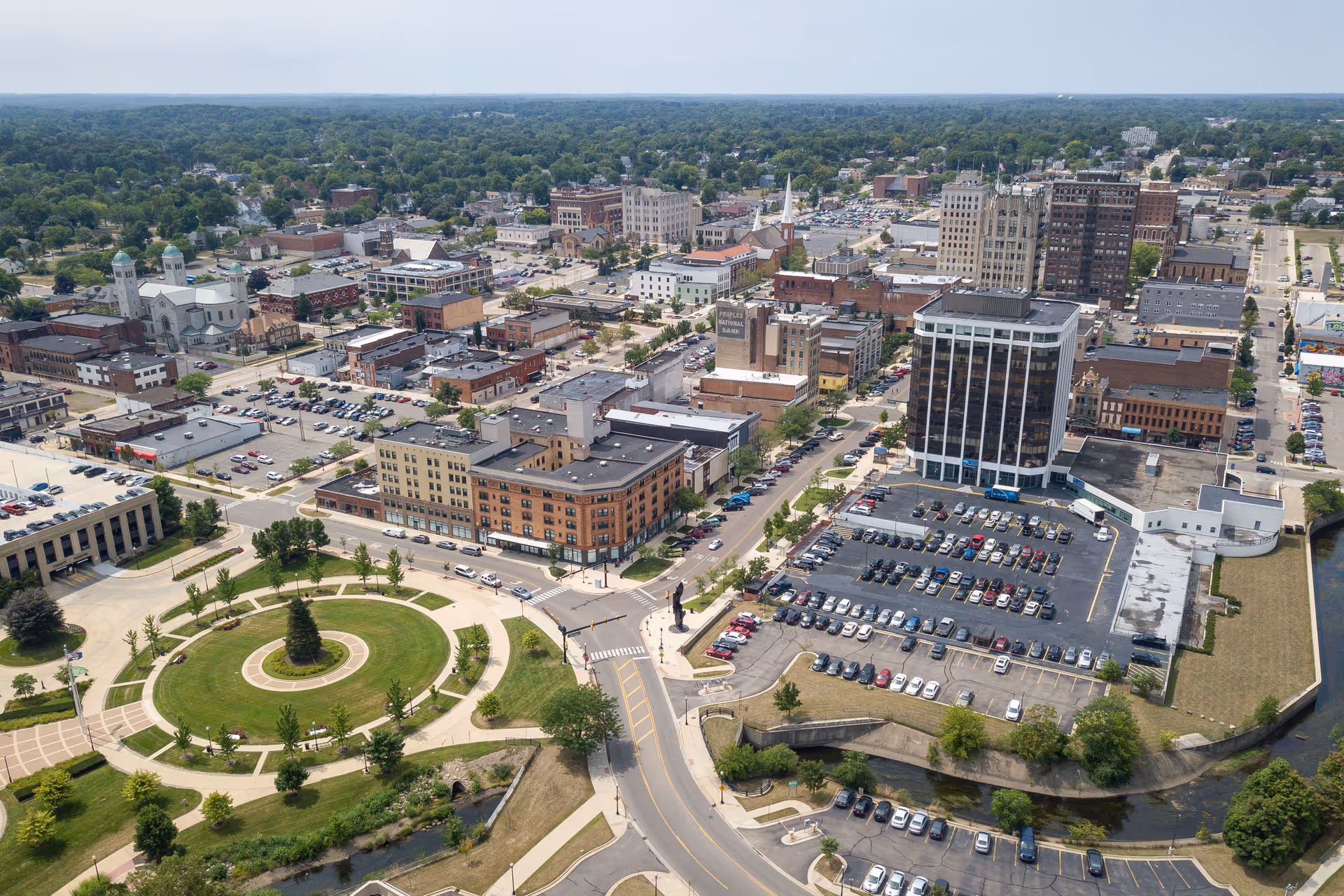 An aerial view of a downtown town with a circular park along a riverfront.