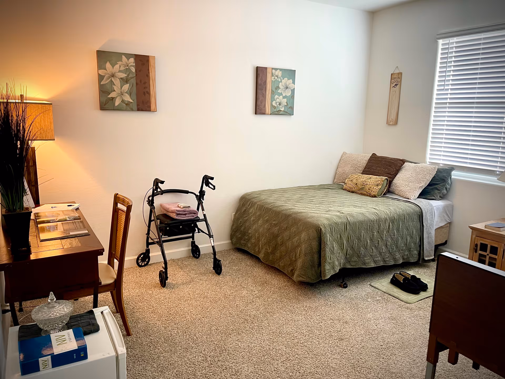 Senior living bedroom with a bed, walker, desk, and window blinds under warm lighting.