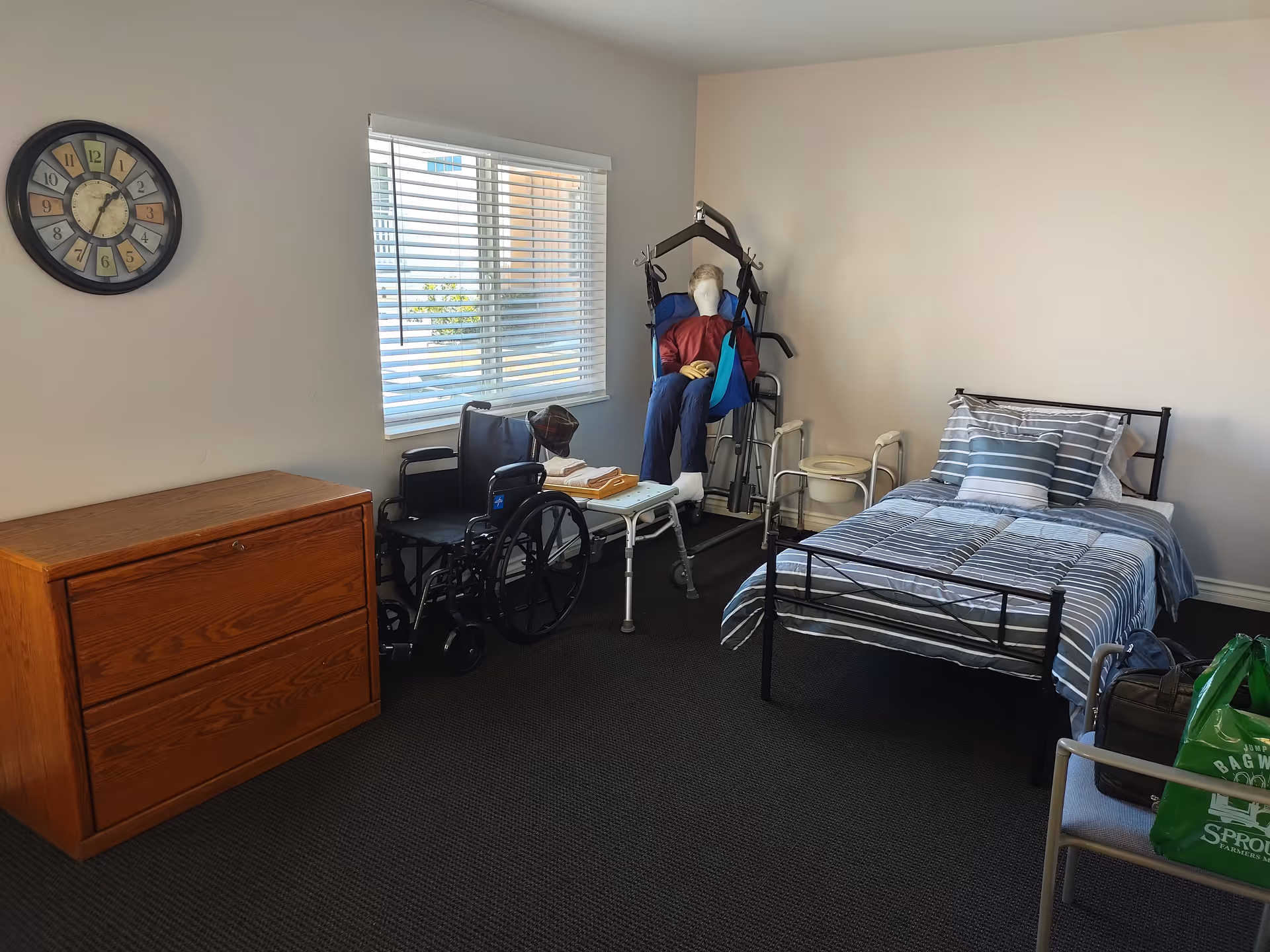 Senior home bedroom featuring a striped bed, dresser, wheelchair, and medical equipment near a window.